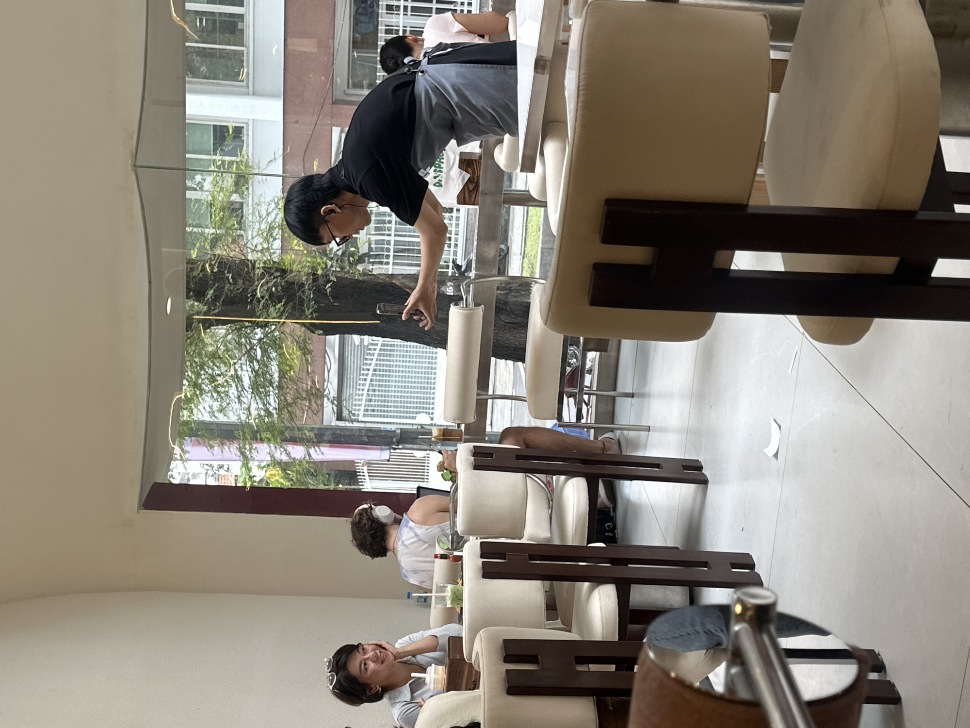 The image displays an interior view of a contemporary café or restaurant with large glass windows looking out onto a street in Thủ Đức, Vietnam, as indicated by a faintly visible "THỦ ĐỨC" sign on an exterior building. The interior features light-colored walls, white tiled flooring, and modern furnishings comprising beige upholstered chairs with dark frames and light-colored tabletops.
Four individuals are present. In the left foreground, a woman with short dark hair and sunglasses perched on her head is seated, smiling with her chin resting on her hand, beside a table holding a light-colored beverage. Further back, a person with curly brown hair and white over-ear headphones is seated, focused on a laptop on their table, accompanied by a drink and a food item. In the right middle ground, a male staff member, wearing a black shirt, glasses, wired earphones, and a gray apron, extends his right arm, holding a smartphone vertically, appearing to photograph or video-record the area in front of him. In the right background, a woman with dark hair is partially visible from behind, seated at a table. A small, crumpled piece of white paper lies on the floor in the center foreground. The exterior visible through the window includes green trees and parts of other buildings.