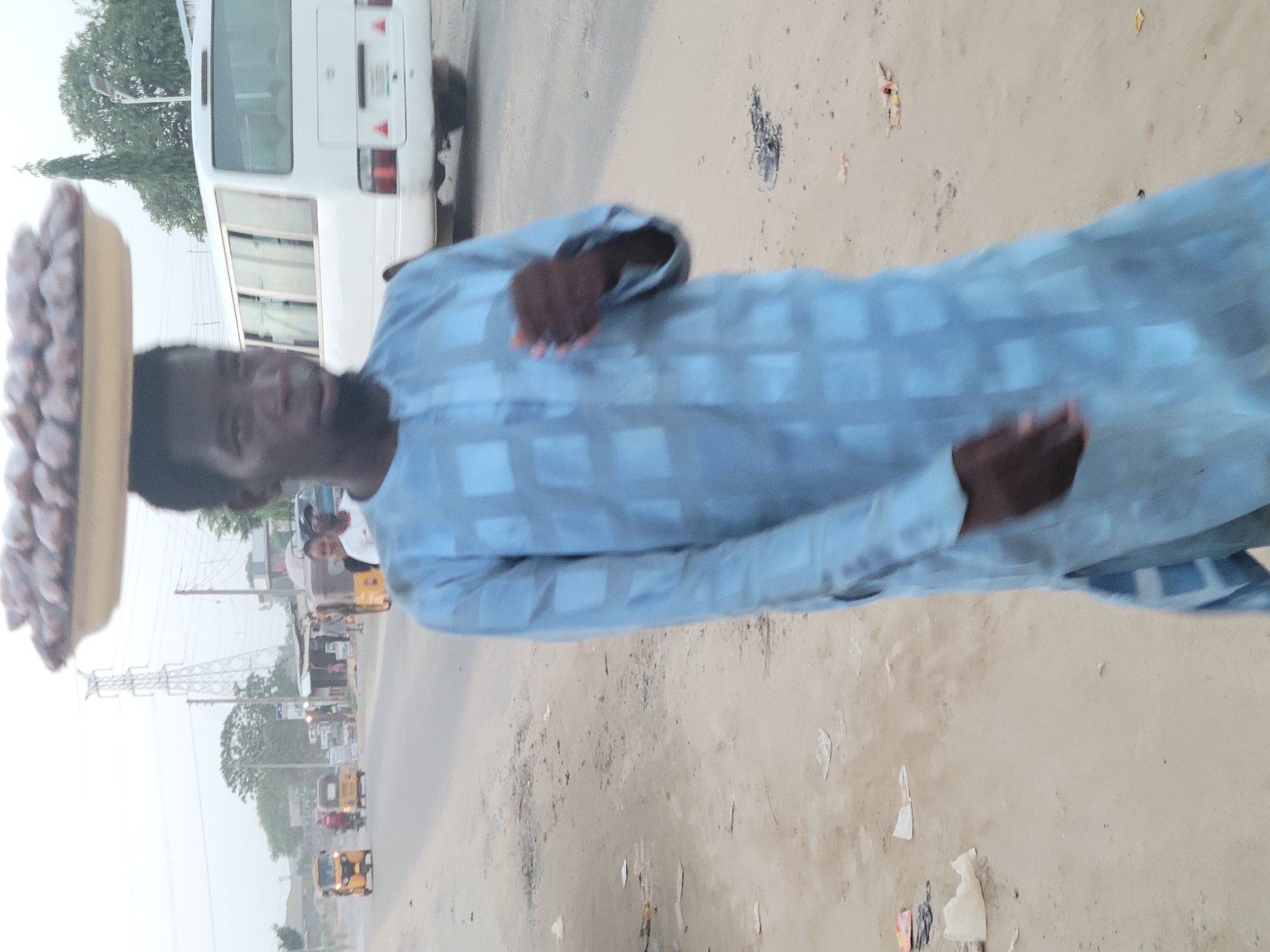 In Madalla, Nigeria, an individual wearing a blue patterned shirt carries a tray of goods on their head. The surroundings consist of a road with a bus to the left and a residential street with several 'Keke Napep' (auto rickshaws) in the distance.