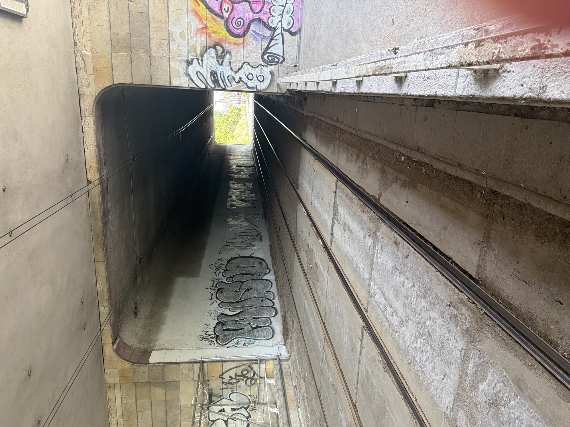 The image depicts a concrete underpass with two sets of train tracks running through it. The underpass is illuminated by natural light from the opening at the far end. The walls of the underpass are covered in graffiti. On the left wall, there is a large piece of graffiti in grey bubble letters that appears to say "PAISID". There are other graffiti tags visible on both walls. The train tracks are weathered and appear to be disused, with debris and dirt accumulated around them. The sky and trees are visible at the end of the underpass, suggesting it leads to an outdoor area. The scene is devoid of people or any signs of recent activity.