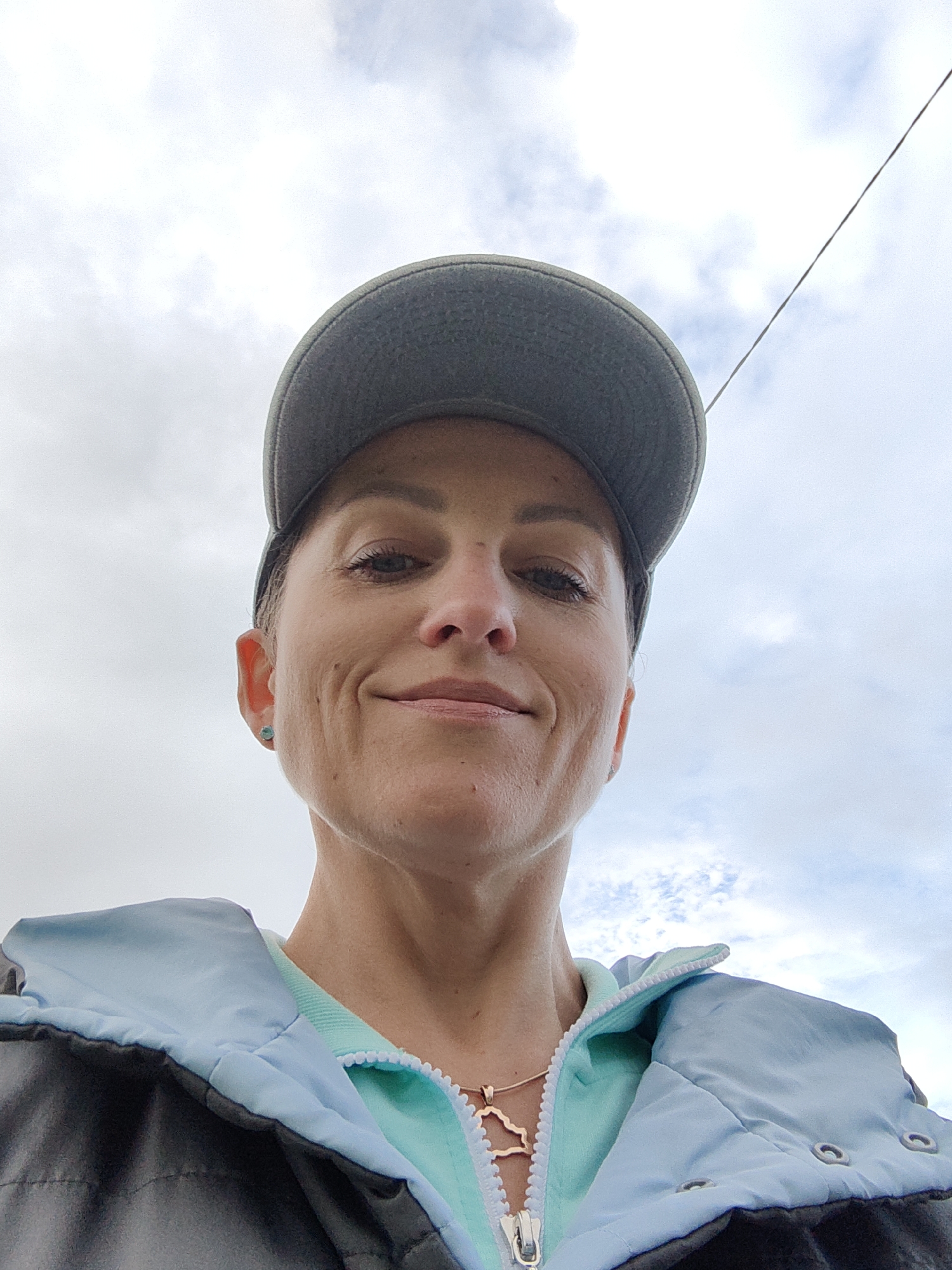 The image shows a low-angle, close-up, selfie-style photograph of a woman wearing a grey baseball cap, a light blue and grey jacket, and a mint green top. She is also wearing a necklace with a gold pendant shaped like the outline of the country of Belarus. The background is an overcast sky with scattered clouds. There is a thin wire visible in the upper right portion of the image. The woman is looking directly at the camera and smiling slightly. There are no specific landmarks visible to indicate the location within Myadzyel, Belarus.