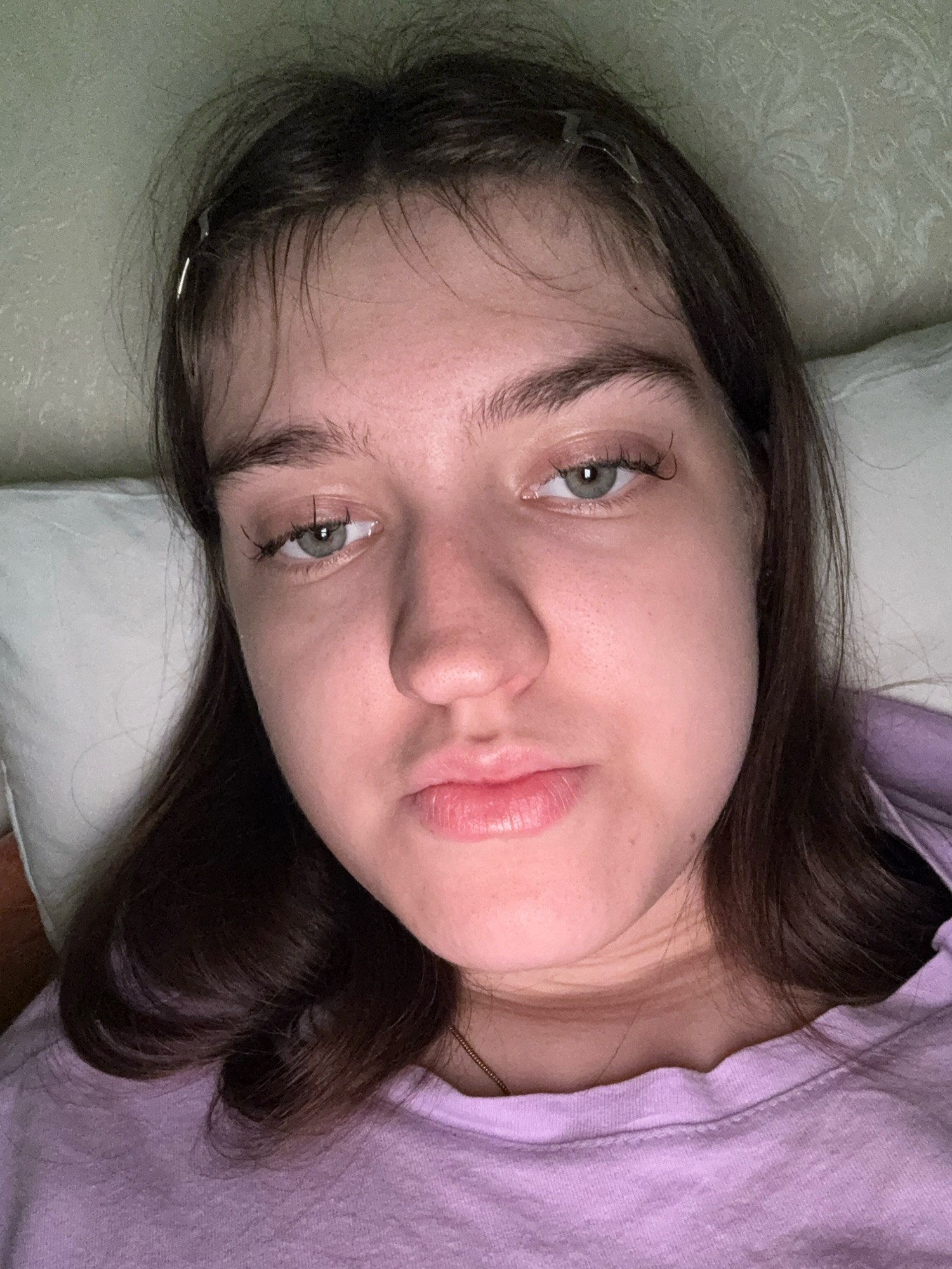 A medium shot, eye-level, head-and-shoulders selfie of a young woman with brown hair and light-colored eyes, is taken indoors. The woman is lying down, with her head resting on a white pillow. She is wearing a light purple sweatshirt. Her hair is loose and falls around her shoulders. She has a neutral expression, with her lips slightly parted. The background is blurred, but appears to be a wall with a textured pattern. The lighting is soft and diffused, suggesting an indoor setting such as a bedroom. The location is unknown, as no specific landmarks or contextual clues are visible in the image. However, the image was taken in Zavolzhye, Russia.