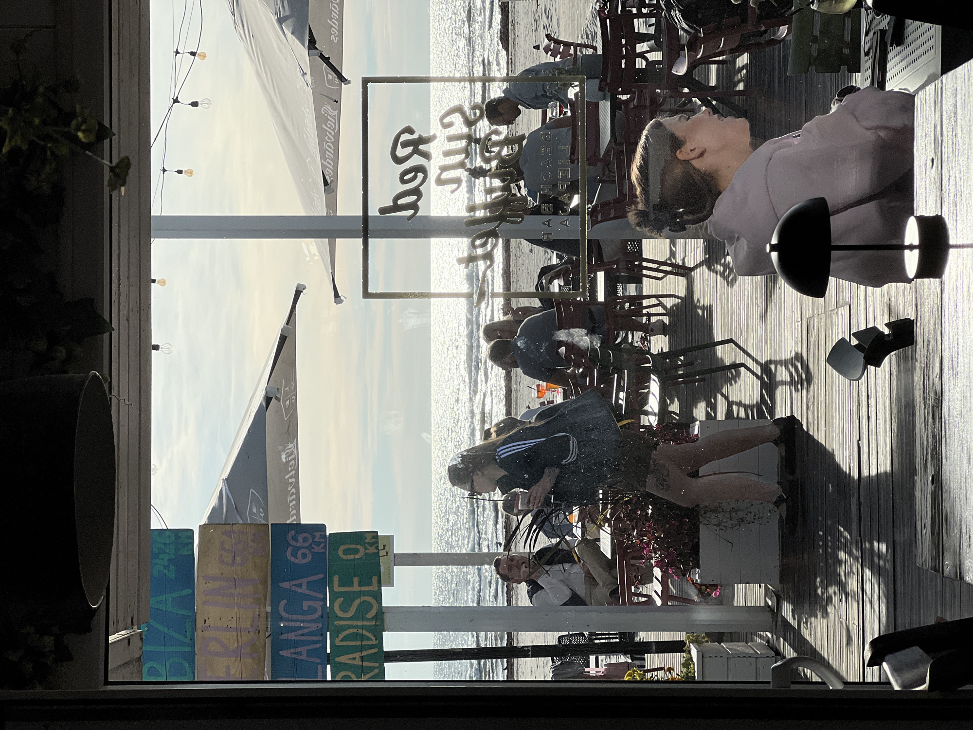 The image captures a sunlit outdoor seating area of a beach bar, likely located in Liepaja, Latvia, given the signs pointing to various destinations. In the foreground, a woman in a light pink hoodie is seated at a table, with a small black lamp beside her. Through a glass window or door, the scene opens up to a view of the Baltic Sea. Several wooden signs are stacked vertically on a pole, indicating distances to "BIZA 2426 KM", "BERLIN 656 KM", "LANGA 66 KM", and "PARADISE 0 KM". People are seated at tables on a wooden deck, some facing the sea, others engaged in conversation. Beach umbrellas provide shade. The overall atmosphere is relaxed and summery.