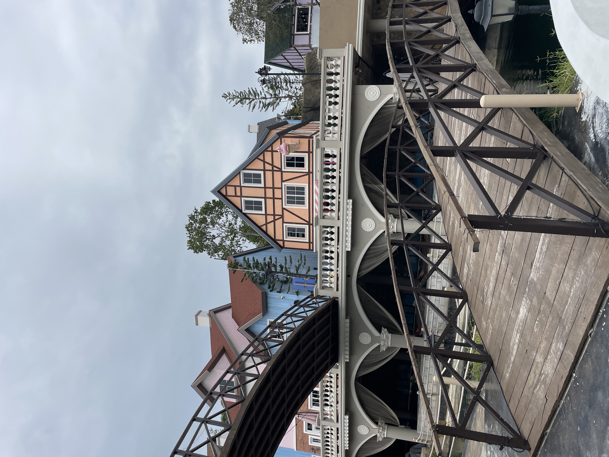 The image shows a scenic view of a European-style village with colorful buildings and bridges in Pattaya City, Thailand. In the foreground, a wooden bridge with dark brown railings spans over a body of water, reflecting the overcast sky. Behind it, another, larger bridge with a similar design arches over a colonnaded structure. The buildings feature a mix of architectural styles, with pastel-colored facades, red-tiled roofs, and prominent window frames. One building stands out with its orange and white half-timbered design, adorned with a pink ice cream cone decoration. White balustrades line the upper level of the structures, adding a touch of elegance. Trees are interspersed among the buildings, and a streetlamp is visible on the right side of the image. The overall scene evokes a quaint, picturesque atmosphere.