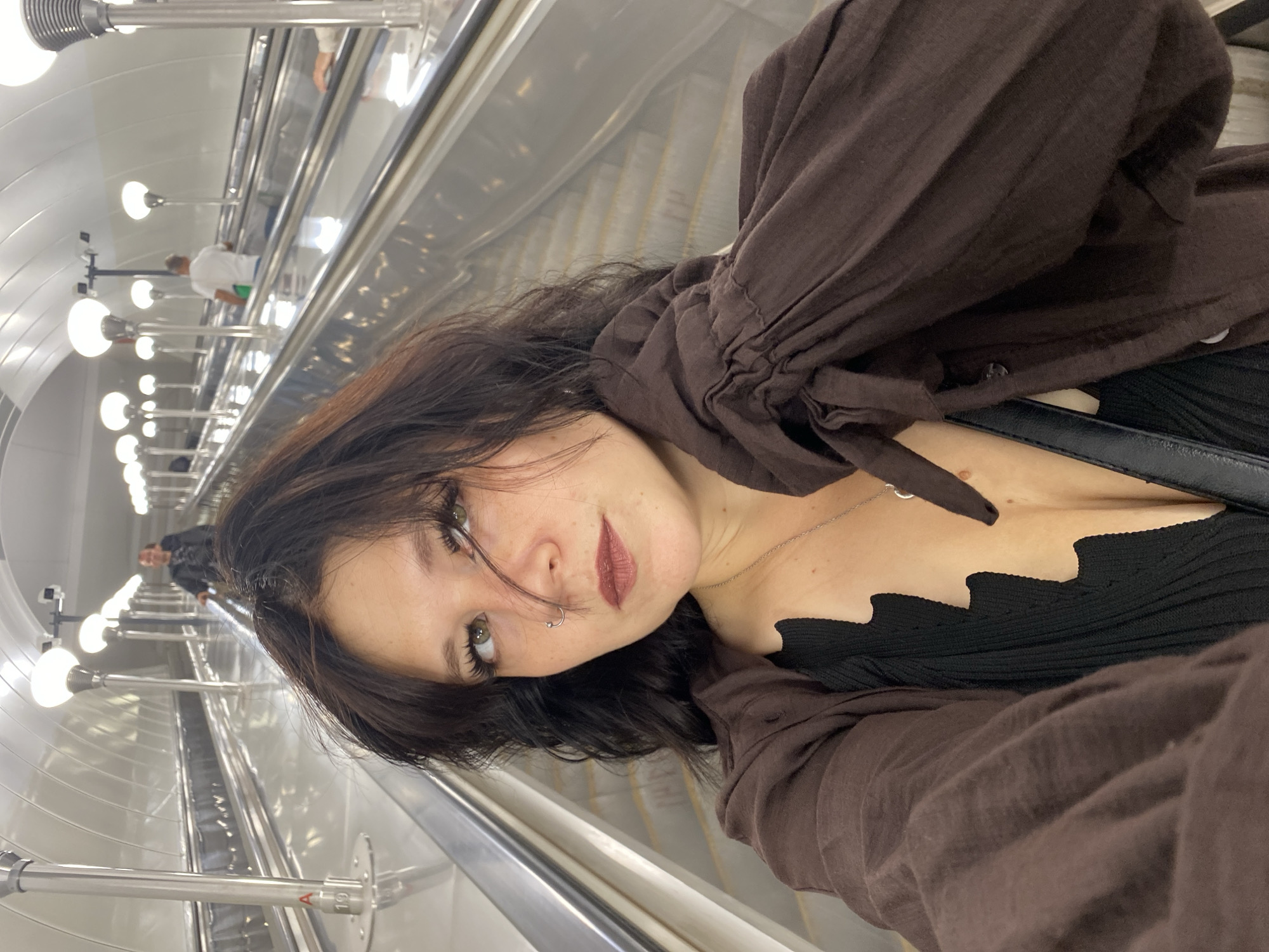 A woman with dark hair, a nose ring, and dark lipstick is taking a selfie on a modern escalator in the Saint Petersburg Metro. She is wearing a brown, ruched-sleeve blouse over a black, V-neck top and a silver necklace. The escalator is metallic and wide, with multiple lanes. The tunnel ceiling is curved and illuminated by rows of modern, dome-shaped lights. In the background, several other people are visible on escalators, including a man in a dark jacket further up and a man in a white t-shirt with a green bag on an adjacent escalator. The overall environment appears clean and well-lit, typical of a Moscow or St. Petersburg metro station.