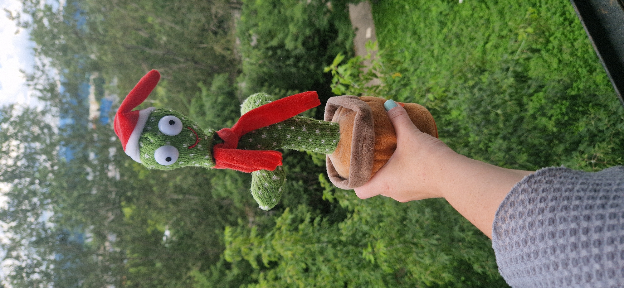 A plush toy cactus wearing a red Santa hat and a red scarf is held by a person's left hand in front of a blurred background of green trees and some distant buildings. The cactus is green with white dots and has large googly eyes and a smiling mouth. Its arms are short and stubby. It is potted in a brown plush pot. The person's hand is light-skinned, and they are wearing a gray knitted sweater. The person's fingernails are painted a light blue color. The background suggests an outdoor setting, possibly a balcony or window overlooking a natural area within a city.