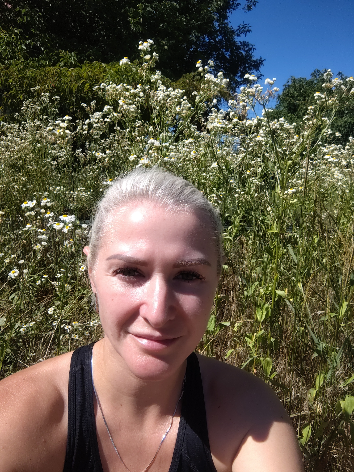 This is a selfie of a woman with blonde hair and fair skin. She is wearing a black tank top and a silver necklace. She is smiling and looking at the camera. She is outdoors, with a field of white flowers and green foliage behind her. The sky is blue and clear. The image appears to have been taken in Hostomel, Ukraine.