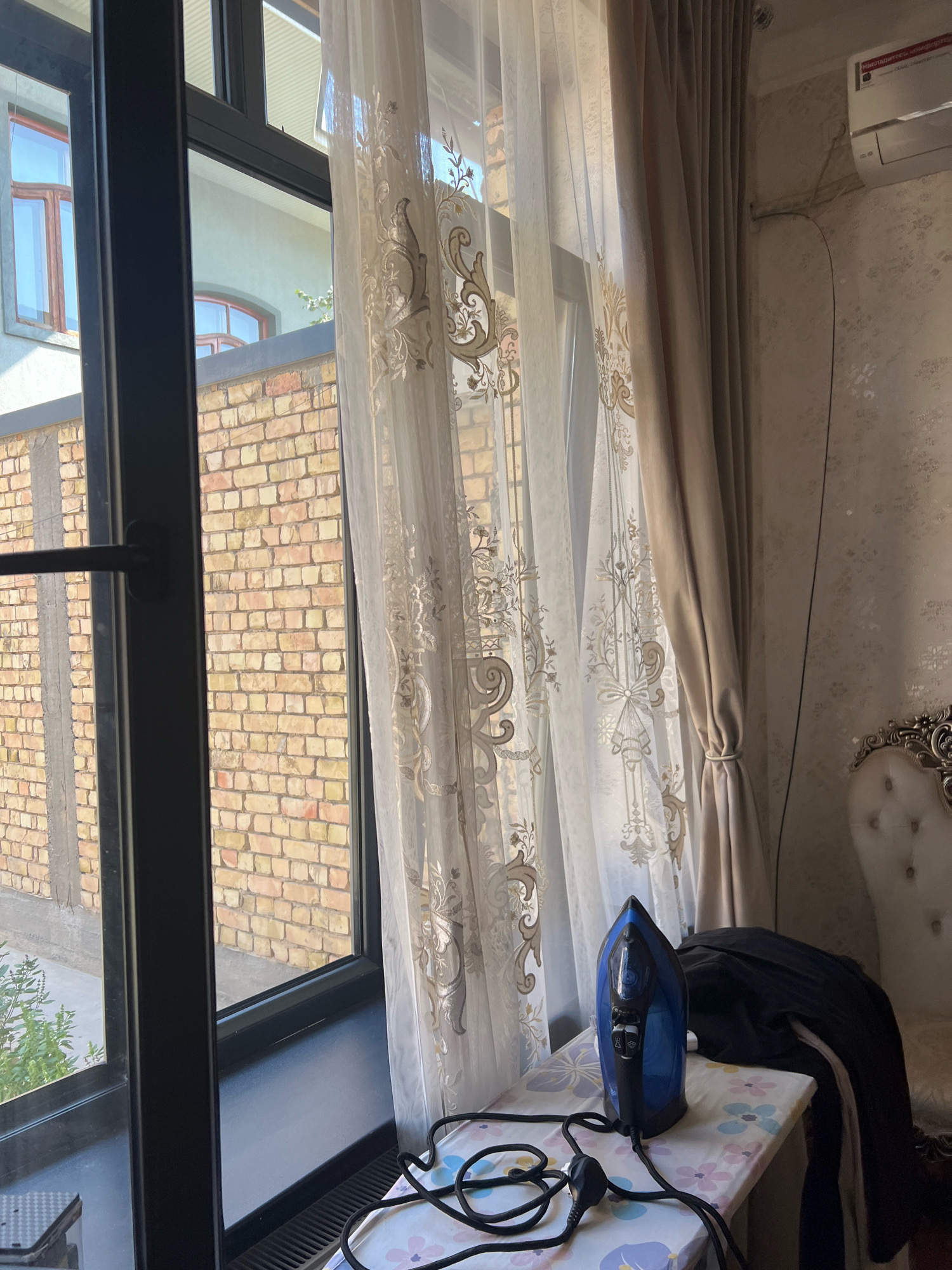 An interior view from an apartment window shows a blue iron on an ironing board next to a draped black garment. The ironing board is positioned in front of a window covered by two layers of curtains, one sheer and white with an embroidered pattern and the other a solid beige. Outside the window, a brick building is visible. To the right of the curtains, a white air conditioning unit is mounted on a textured wall. A portion of an ornate white chair is visible in the lower right corner.