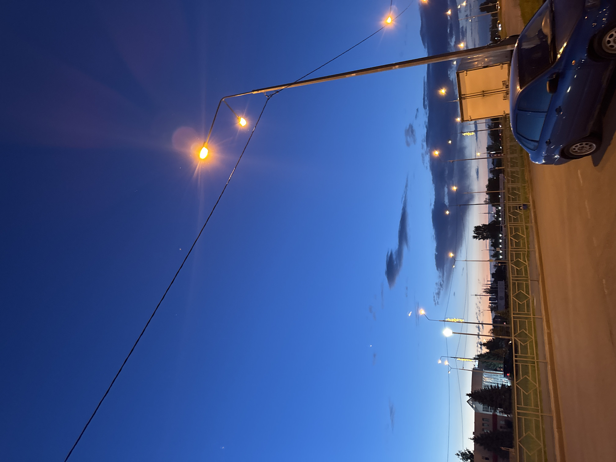The image depicts an urban scene at dusk in an unknown city. The sky is a deep blue, transitioning to lighter hues towards the horizon. Streetlights illuminate the scene, with their warm glow contrasting against the cool sky. A blue car is parked on the side of the road. The road is adjacent to a sidewalk. Buildings and trees are visible in the distance, silhouetted against the twilight sky. A utility pole and wires are also present.