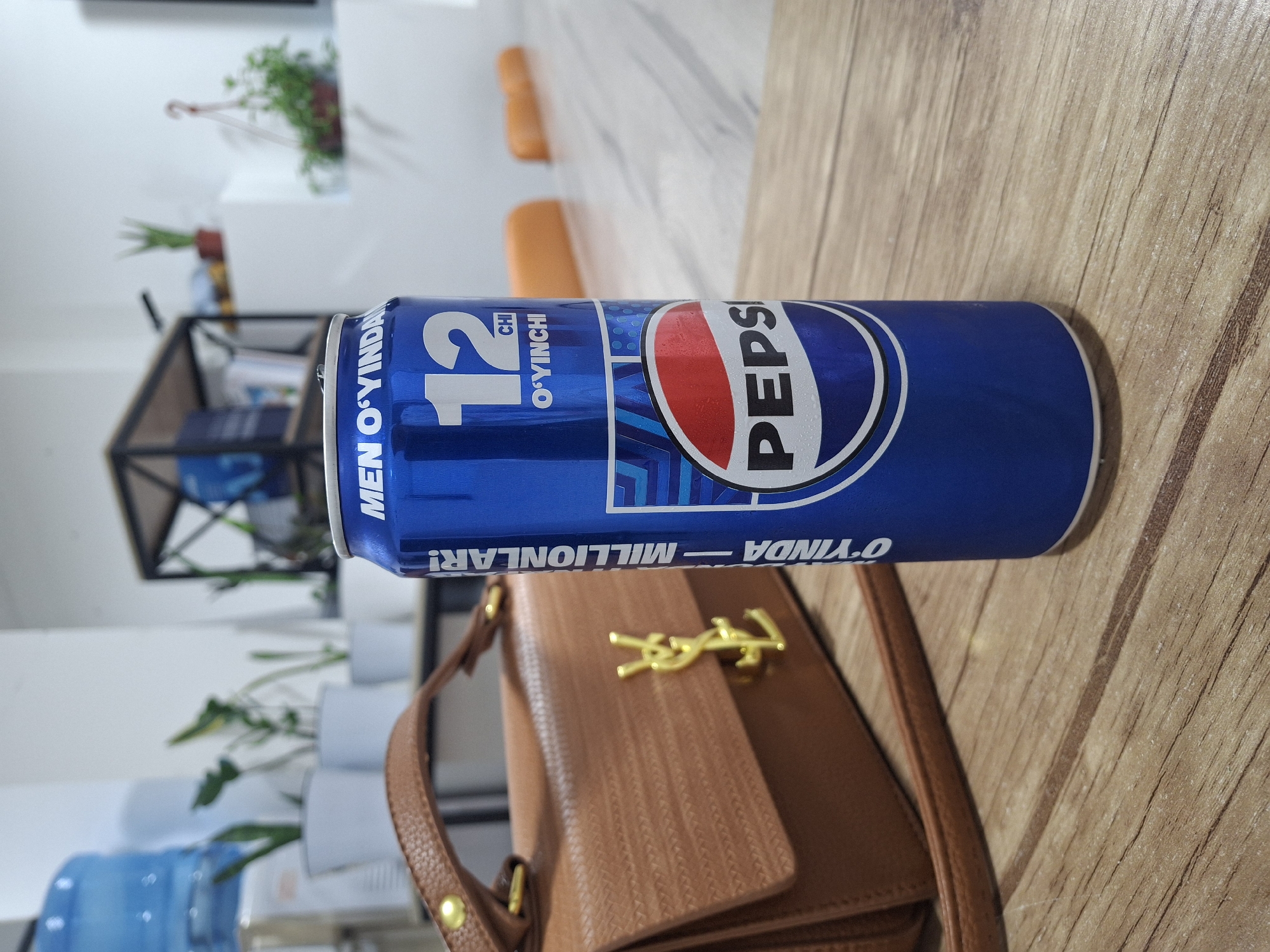 A close-up, eye-level shot focuses on a blue Pepsi can placed on a wooden table. The can is positioned on the right side of the frame and is the primary subject. Text on the can is visible in white and features a prominent "12 CHI O'YINCHI" and the Pepsi logo. To the left of the can, a light brown, textured handbag with gold hardware is partially visible, with its strap extending diagonally across the lower left portion of the image. The background is blurred, showing an office or indoor environment with white walls, potted plants, and shelves with books or binders. Orange chairs are faintly visible in the far background. The lighting appears to be artificial, illuminating the scene from the front and top. The image does not provide any specific geographical markers for Tashkent, Uzbekistan; the description is limited to the observable elements within the photograph.