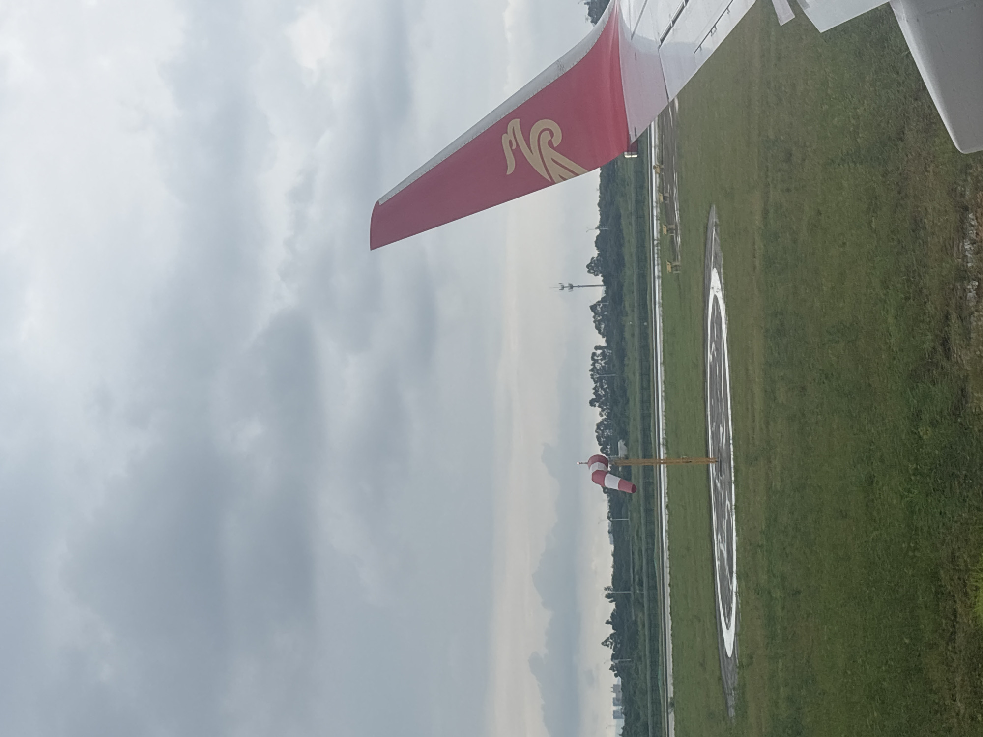 The image shows an airplane wing on the right, with a red tail fin featuring a gold eagle logo. In the background, a windsock with red and white stripes stands on a yellow pole near a circular landing pad marked with white lines. The scene is set on a grassy field with a cloudy sky overhead. Buildings and trees are visible in the distance, suggesting an airport environment in a city, likely Baiyun District, China, given the context provided. The perspective suggests the photo was taken from inside another aircraft or from a window at the airport.