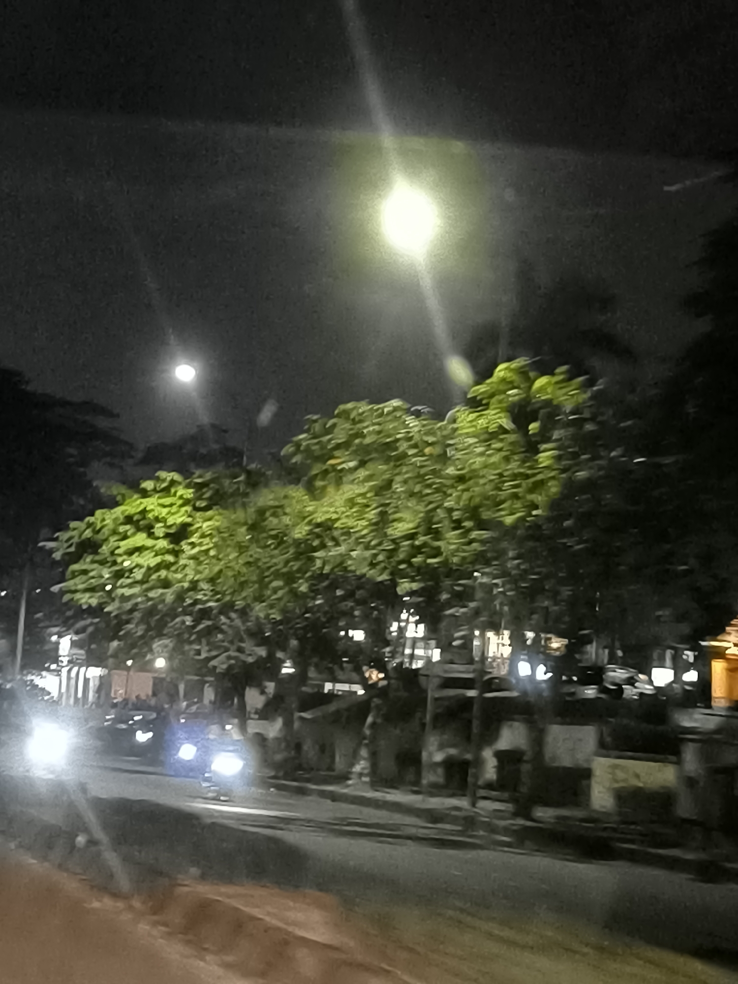 The image depicts a street scene at night in Tangerang, Indonesia. A bright street lamp illuminates the upper portion of the frame, casting a yellow glow. Below, a line of trees with vibrant green foliage stands out against the dark sky. The trees appear to be on the side of a road, where several vehicles are visible. One car in the foreground has its headlights on, casting a bright white light. Other vehicles and buildings are dimly lit in the background. The overall impression is of a typical urban night scene, possibly on a busy street.