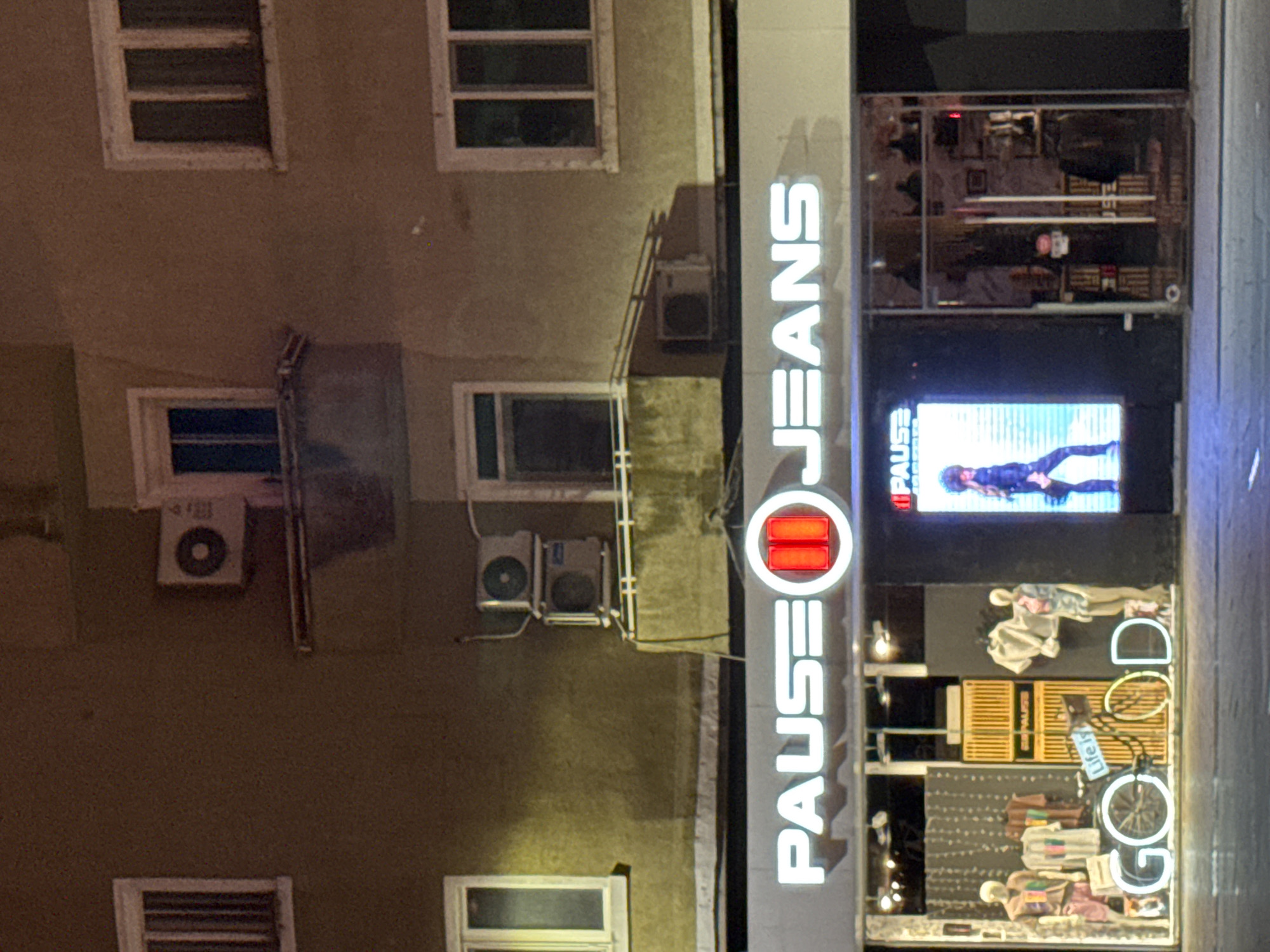 The image shows a nighttime scene of the "PAUSE JEANS" store located in Varna, Bulgaria. The store's illuminated signage is prominently displayed, and the building's facade, including windows and air conditioning units, is visible. The storefront features a display with mannequins, clothing, and a video screen showcasing a woman in a black outfit. The store is located on a street.