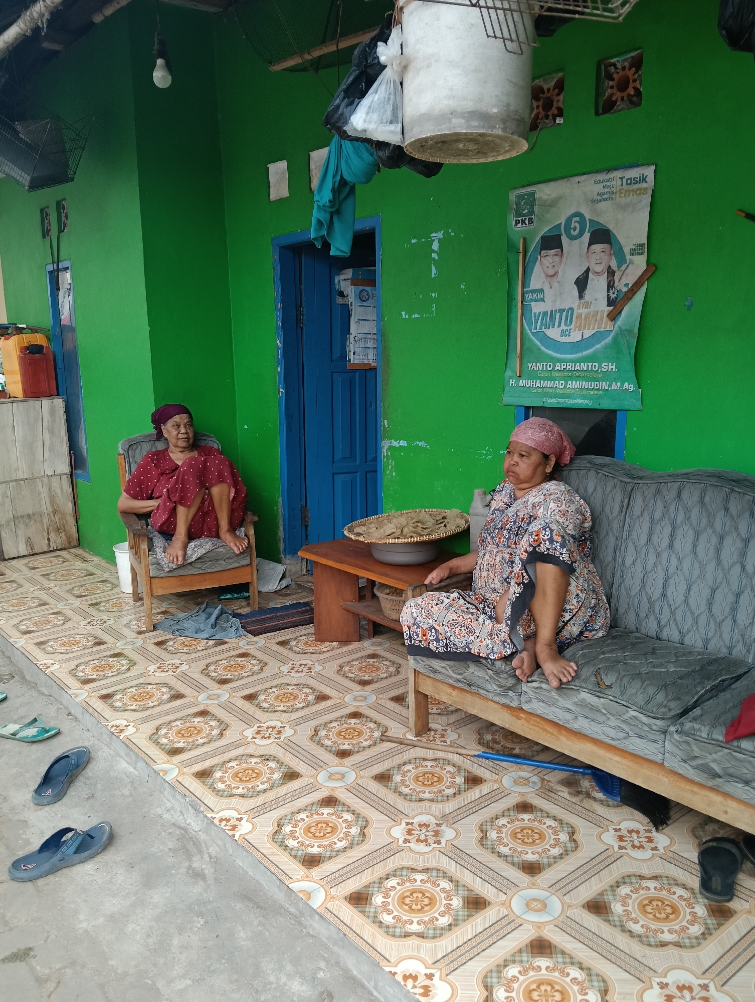 Two elderly Indonesian women are seated outdoors in front of a bright green building in Tasikmalaya, Indonesia. The woman on the left sits in a wooden armchair, wearing a red polka dot dress and a dark headscarf. Her legs are crossed, and her feet are bare. To her right, a wooden table holds a woven basket filled with crackers. The woman on the right is seated on a worn, grey couch, dressed in a patterned long-sleeved dress and a light pink headscarf. Her legs are extended, and her feet are bare. The building behind them has a blue door, and a political poster is visible on the wall to the right. The ground is tiled with a repeating floral pattern. Several pairs of flip-flops are scattered on the concrete area in front of the tiled walkway. The scene suggests a humble, everyday setting, likely a residential area in Tasikmalaya.