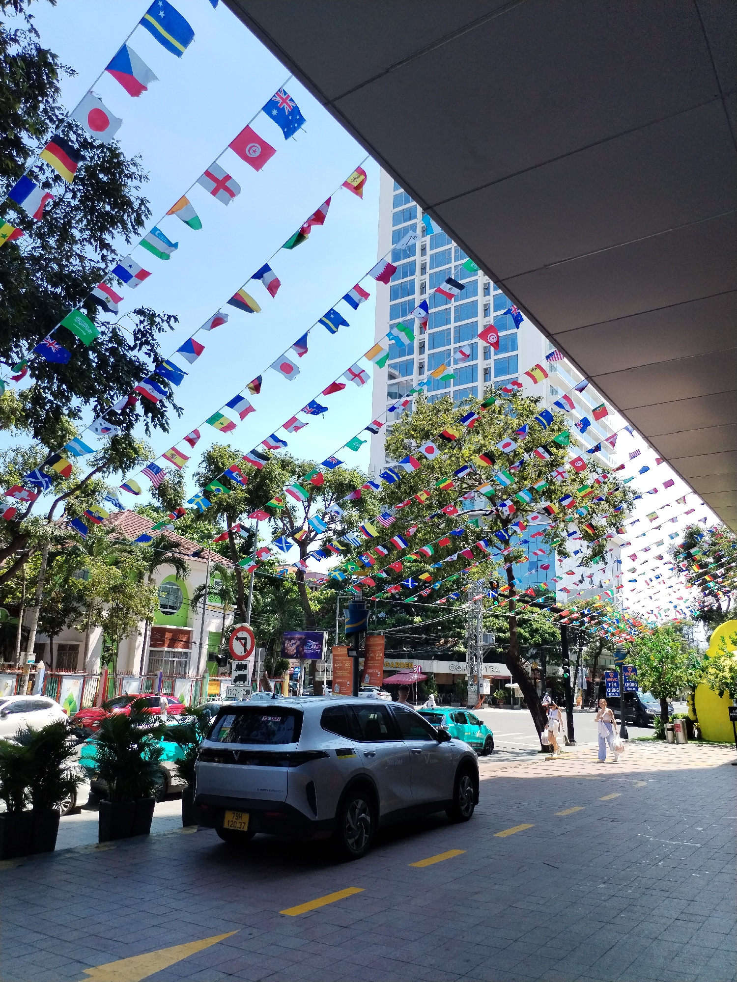 This media file depicts a street scene in Nha Trang Ward, Vietnam, likely during the day. The foreground is dominated by a silver SUV with a license plate reading "79H 120.37". The street is paved with interlocking bricks and marked with yellow directional arrows. In the background, a modern building with many windows stands tall, contrasting with the surrounding trees and smaller, older buildings.
The most prominent decorative element is a vast display of national flags strung across the street, creating a colorful canopy. These flags suggest a festive or international event is being celebrated.
Several people are visible walking along the sidewalk, some carrying shopping bags, indicating a normal day of activity. One person appears to be a taxi driver near the silver SUV. A red car and a teal-colored car are also parked or driving on the street.
A traffic sign with a red circle and a diagonal red line over a black arrow pointing right is visible, along with a traffic light. Signage in Vietnamese is present, including "GARDENS", "COF TEA", and text that appears to be advertising or informational, such as "TIÊM CHÚNG". The time "16:00-17:00" is also visible on a sign, suggesting it is late afternoon. The weather appears to be sunny with a clear blue sky, contributing to the vibrant colors of the flags.