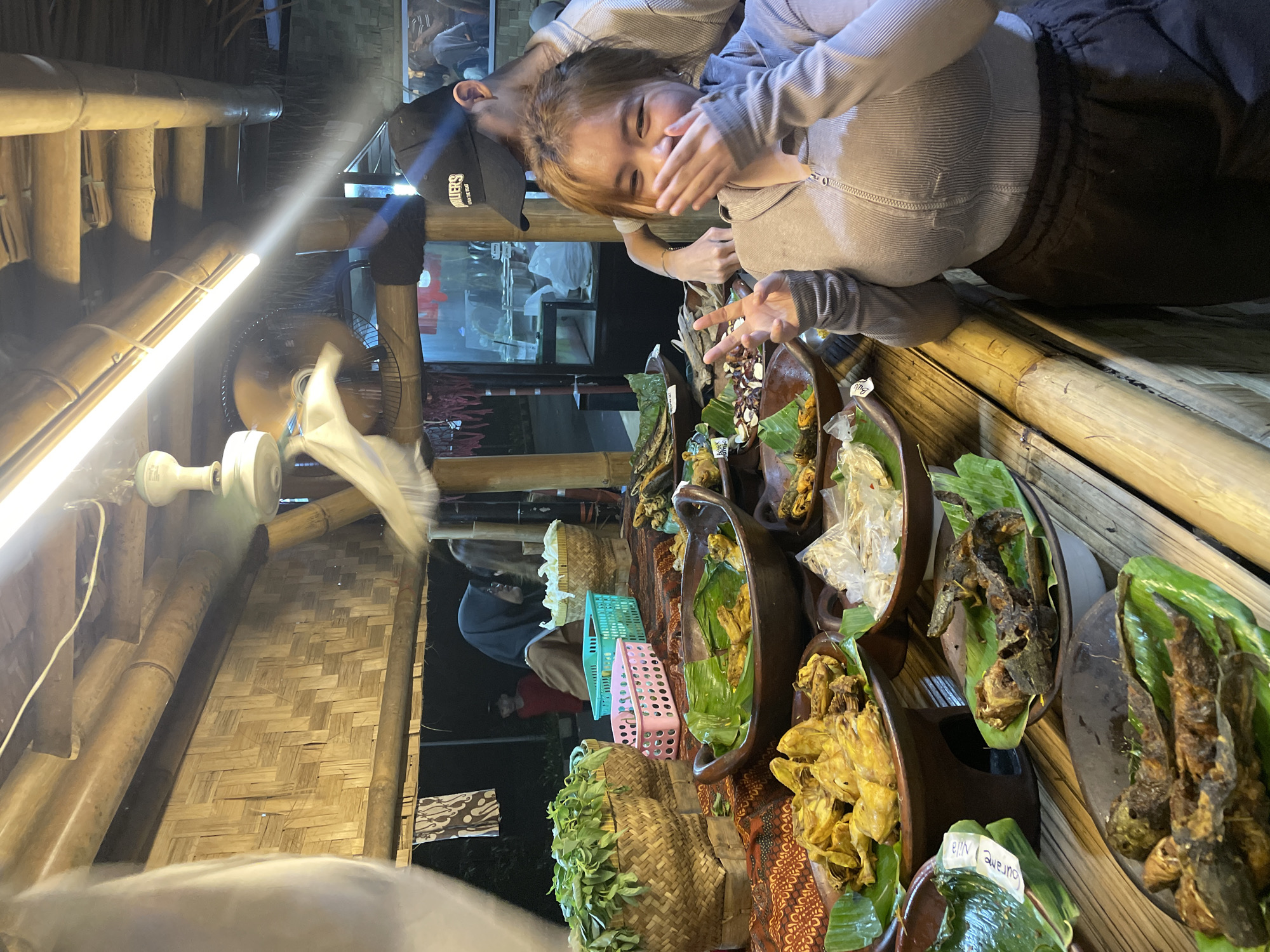 Captured at night, the scene unfolds in North Jakarta, Indonesia. A young woman with light brown hair, clad in a gray long-sleeved shirt and dark pants, stands at a food stall. She covers her mouth with her left hand, while her right hand makes a "peace" sign near the food. To her left, a person wearing a black cap leans over the stall.
The food stall is lined with a variety of dishes, each served on banana leaves and displayed in dark brown, boat-shaped containers. The dishes include what appears to be fried chicken, fish, and vegetables. Some food items are labeled with tags. Behind the food stall, there are woven baskets, some filled with leafy vegetables, and stacked pastel-colored crates.
The stall itself seems to be constructed from bamboo, with a woven bamboo wall visible in the background. An old-fashioned fan hangs from the ceiling. The overall lighting is warm and dim, highlighting the food and the people at the stall.