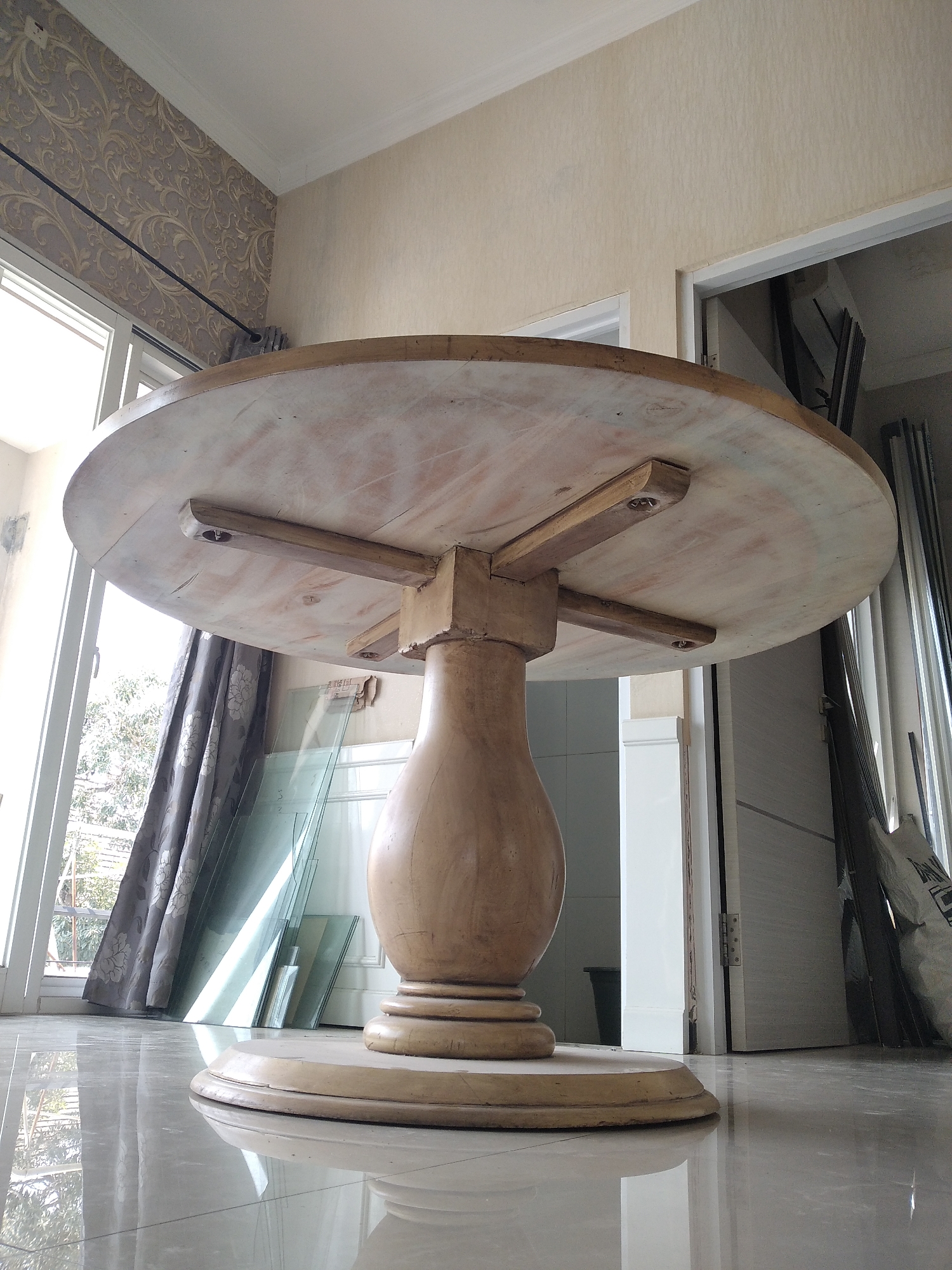 The image shows a low-angle, close-up view of the underside of a light-colored, circular wooden table with a pedestal base. The table has visible wood grain and some areas of discoloration, suggesting it may be unfinished or aged. Four wooden supports are visible beneath the tabletop, attached with screws. The pedestal base is bulbous and tapers towards a wider, circular foot.
The table is situated indoors, on a highly reflective tiled floor that mirrors the table and its surroundings. To the left of the table, a large window with white frames reveals greenery and trees outside, along with a gray curtain with a floral pattern. In the background, to the right of the table, is an open doorway leading to another room. Various objects are visible in the background, including what appear to be sheets of glass leaning against the wall, stacked construction materials, and a closed door. The walls are painted in a light, neutral color, with a section of patterned wallpaper above the window on the left.
The location is described as being in Kaliwates, Indonesia. The scene appears to be a room within a residential or commercial building, possibly under construction or renovation, given the presence of building materials and sheets of glass. There are no people or animals present in the image.