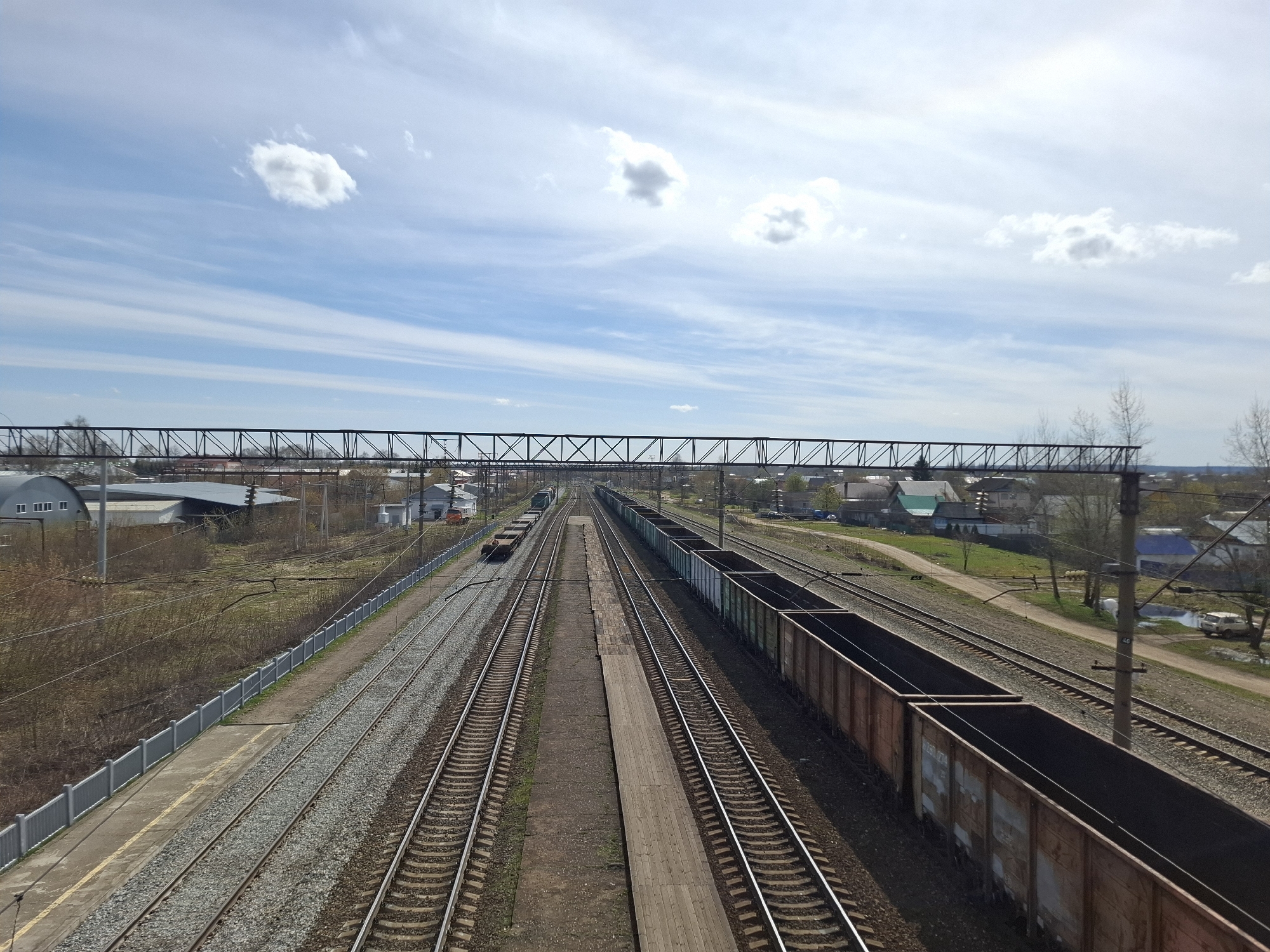 The image shows a train station area in Амзя, Russia. There are multiple railway tracks running parallel, with trains visible on some of them. Various buildings, houses, and vegetation are present in the background, suggesting a mix of residential and industrial areas. The sky is blue with scattered clouds. Overhead, a metal structure spans across the tracks, likely supporting electrical lines or signals. A long platform runs between two sets of tracks. The photo appears to have been taken from an elevated position, providing a wide perspective of the station and its surroundings.