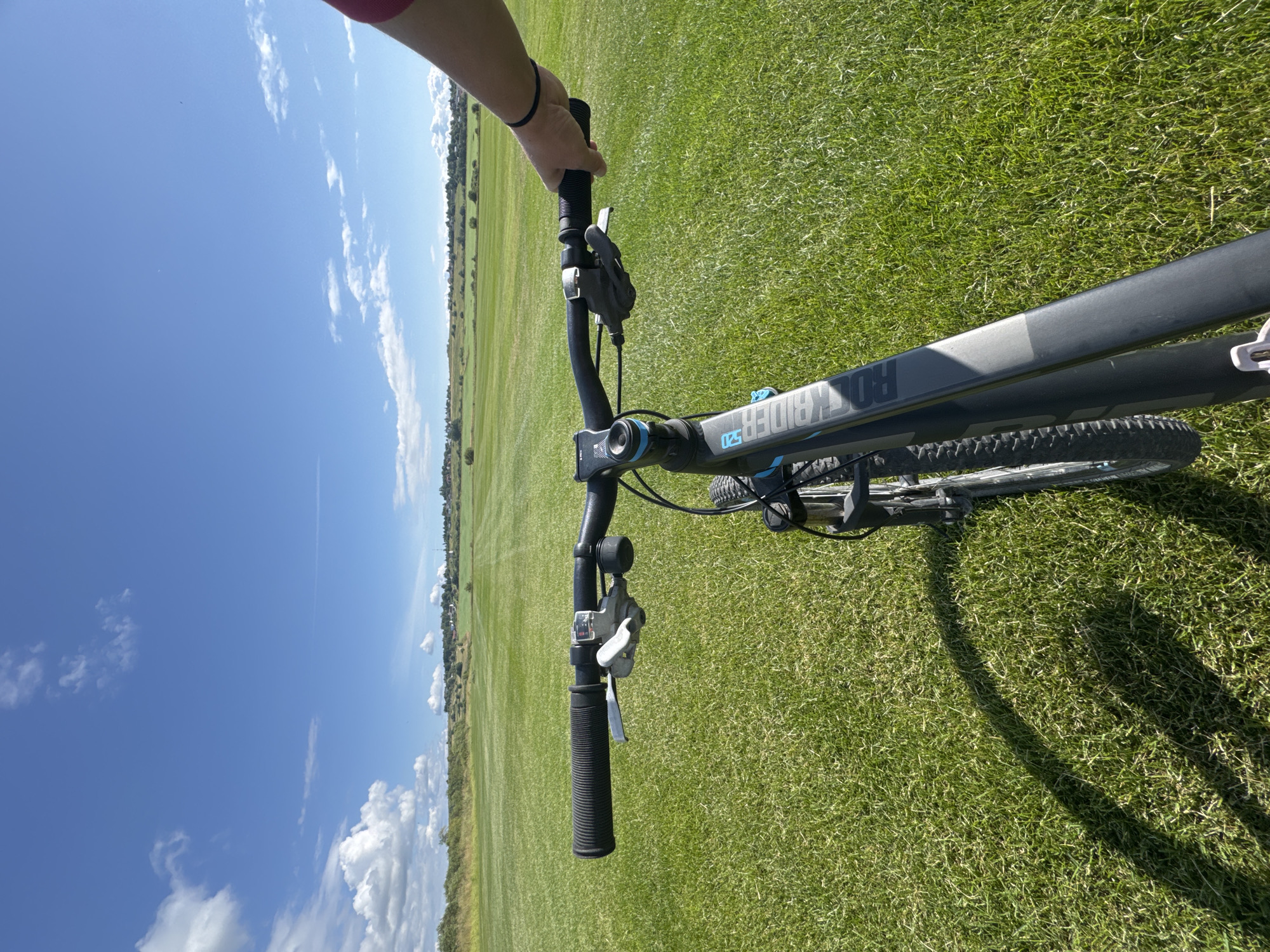 A first-person perspective shot of a person riding a black mountain bike on a vast, well-maintained green lawn under a clear blue sky with scattered white clouds. The handlebars of the bicycle are in the foreground, with the rider's left hand gripping the left handlebar and their right hand holding the right handlebar. The bike's frame is black with blue accents and the brand name "ROCKRIDER" is visible. The lawn extends into the distance, with rows of mown grass visible. In the far distance, a line of trees and some buildings can be seen along a gentle slope. The scene is well-lit, indicating daytime. The location is the city of Kempelevo, Russia.