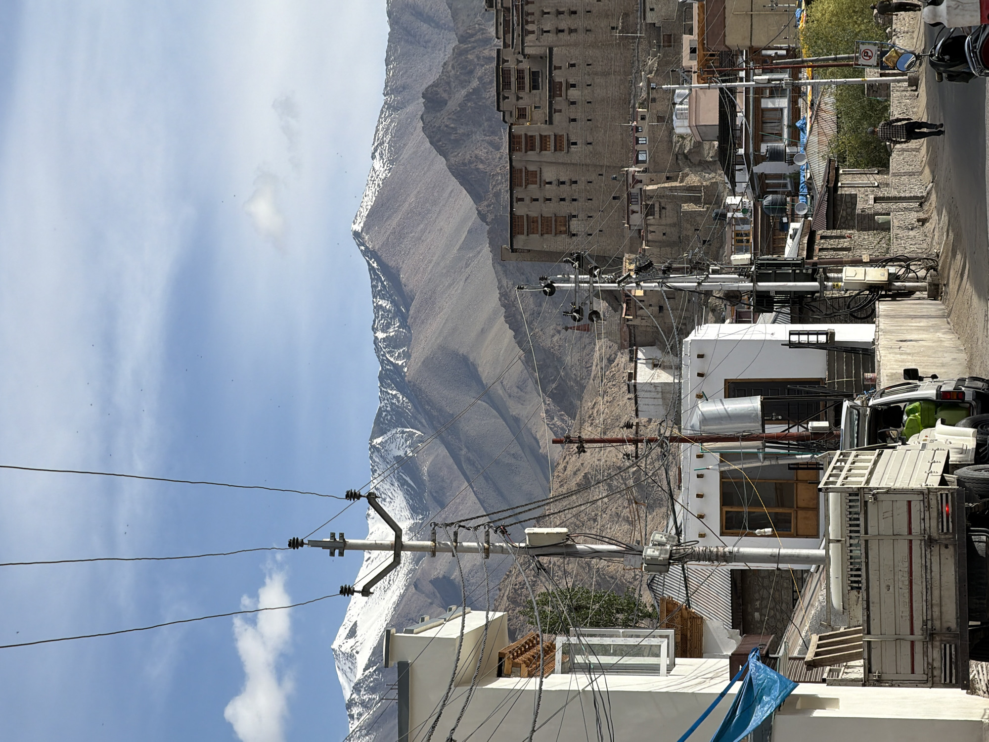 The image depicts a street scene in Leh, India, with the majestic snow-capped Stok Kangri mountain range forming a prominent backdrop. In the foreground, a white truck with a flatbed is parked on the right side of the road. The truck has a visible "Mahindra" logo on its side. To the left of the truck, a white building with a blue tarp is visible. Numerous power lines crisscross the scene, suspended from utility poles, contributing to a sense of urban infrastructure against the natural landscape.
Further down the street, several other buildings of varying sizes and architectural styles are visible, including a large, multi-story structure with numerous windows that appears to be a historical building or monastery, possibly the Leh Palace or a similar landmark, given its scale and construction. People are seen walking along the street, suggesting normal daily activity. The overall environment appears to be a mix of residential and commercial buildings, characteristic of a town. The sky is partly cloudy, with a clear blue sky dominating the upper portion of the image. The light suggests it is daytime, likely mid-morning or afternoon.