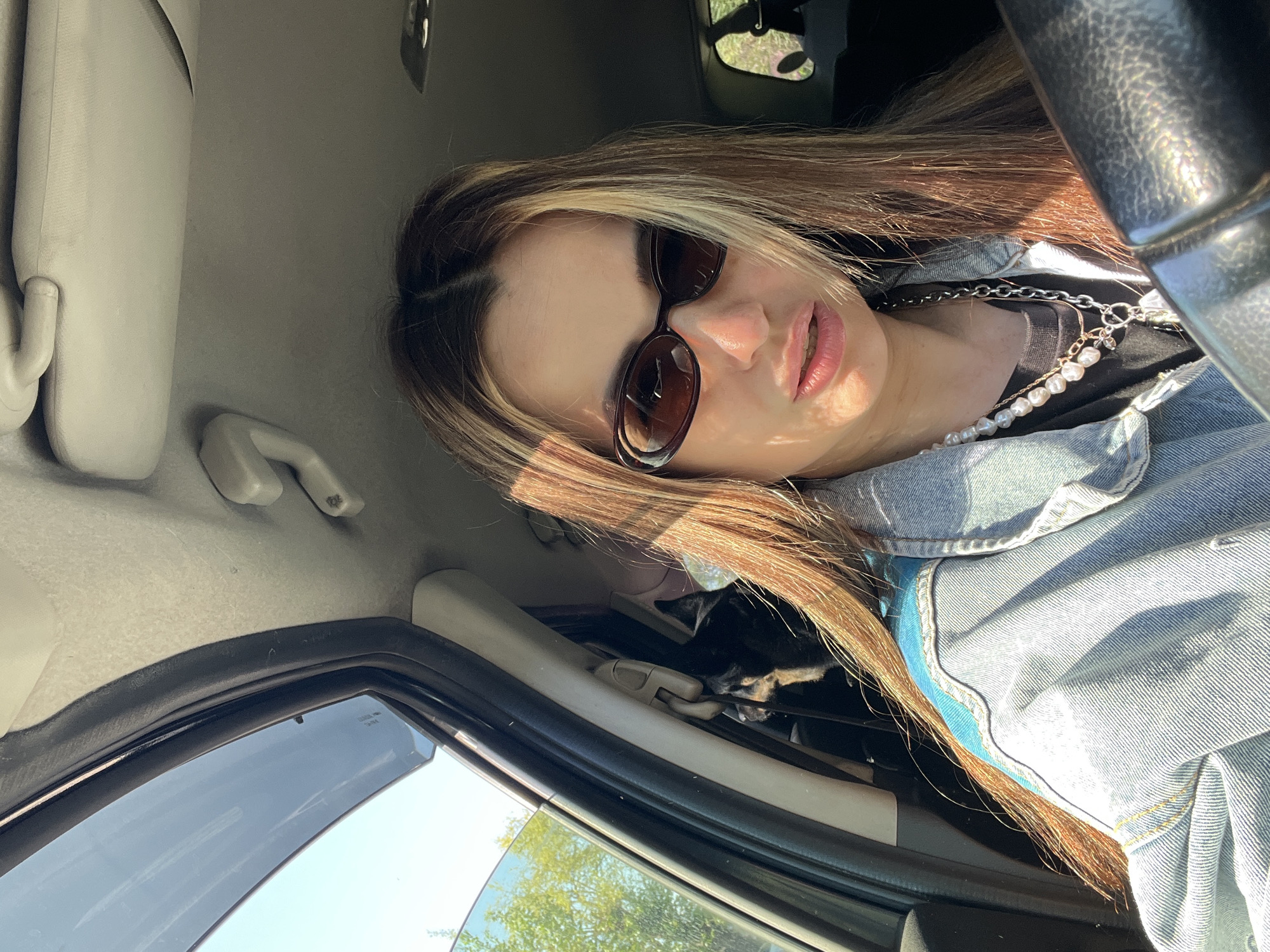 A young woman with long, light brown hair and sunglasses is seated inside a car in an unknown location in an unknown city. She is wearing a blue denim jacket over a black shirt and a pearl necklace. The interior of the car is light gray. Light is entering through the car window. The steering wheel is visible in the upper right corner.