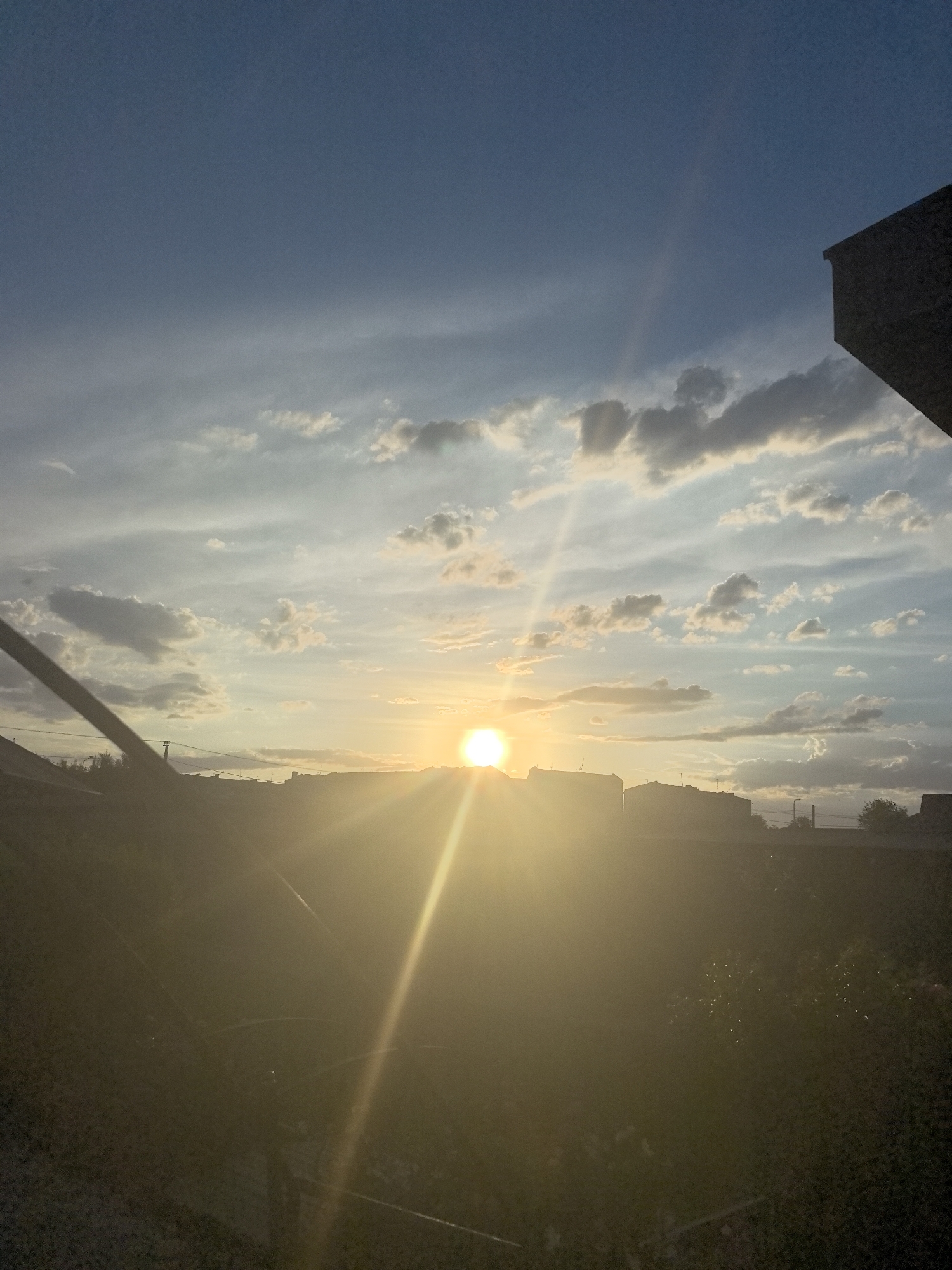 The image captures a sunset over a residential area, possibly in Taldykorgan, Kazakhstan. The sun is a bright, golden orb positioned low on the horizon, casting a warm glow and creating lens flare that streaks across the scene. Wispy clouds, tinged with yellow and orange hues from the setting sun, are scattered across a vast, blue sky.
In the foreground, silhouetted against the bright sky and the sun's glare, are the rooftops of several buildings, suggesting an urban or suburban environment. A dark, diagonal line, likely a wire or a part of a structure, cuts across the left side of the image, partially obscuring the view. The lower portion of the image is very dark and appears to show vegetation, possibly a garden or bushes, also silhouetted and difficult to discern in detail due to the low light and glare.
The overall mood is serene and peaceful, characteristic of a sunset. The lighting suggests it is late afternoon or early evening. There is no visible text in the image. The presence of buildings and what appears to be a fence or wall in the foreground, along with the composition suggesting a view from an elevated or sheltered position, provides context for the scene.