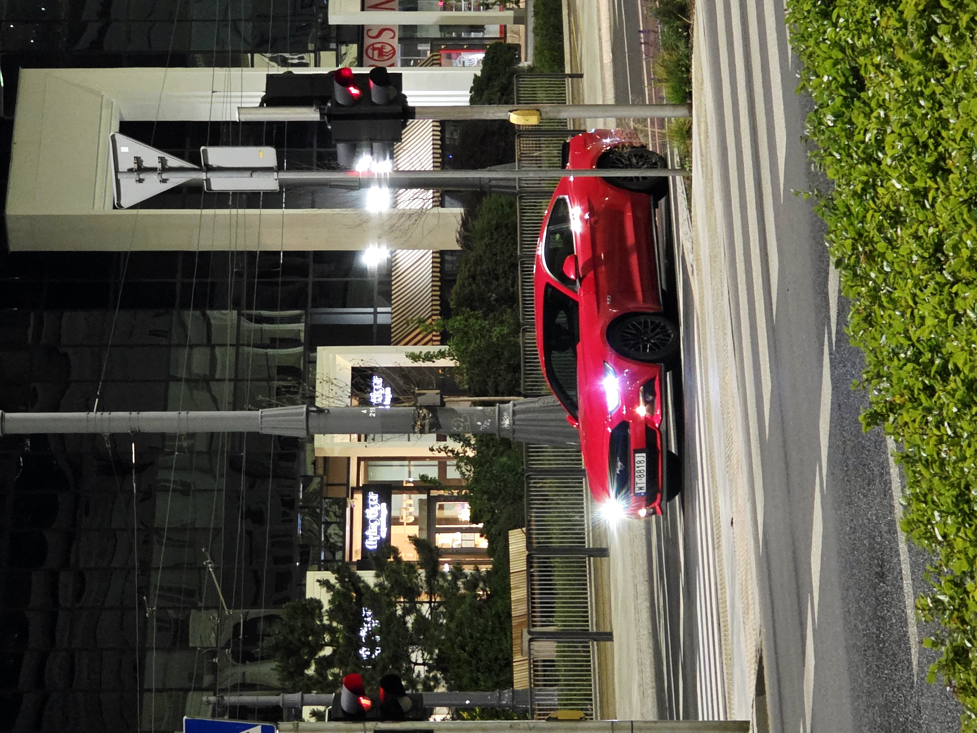 A red Ford Mustang is stopped at a pedestrian crossing on a city street at night. The car's headlights are on and shining brightly. In the background, a modern building with large glass windows reflects the city lights. A traffic light is visible, showing a red signal. The street is paved, and there are white stripes marking the pedestrian crossing. Green bushes are in the foreground. The license plate on the car reads "WT 8818J". This scene appears to be in Warsaw, Poland, based on the license plate format and the architecture. There are no people visible in the image.