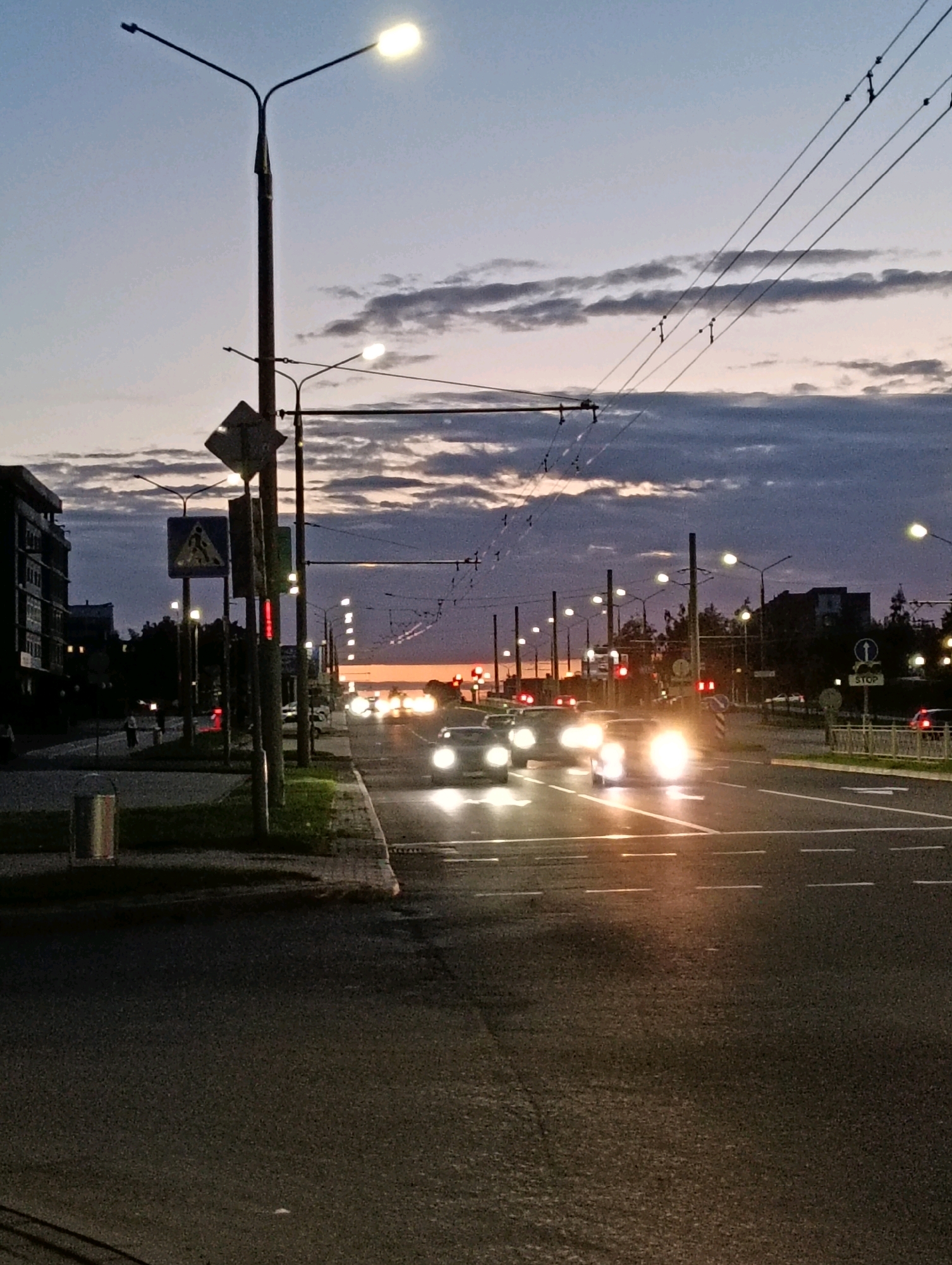 This image shows a street scene at dusk in Hrodna, Belarus. The sky is a gradient of pale blue and grey, with streaks of orange and pink near the horizon, indicating the sun has recently set. Several cars are driving on the road, their headlights illuminated, casting bright reflections on the wet asphalt. Overhead, a network of power lines and streetlights crisscross the sky, with some lights already on. The street is lined with trees and buildings, some of which have illuminated windows. On the sidewalk, there are trash cans, traffic signs, and a few pedestrians walking. The overall impression is of a busy urban environment transitioning from day to night.