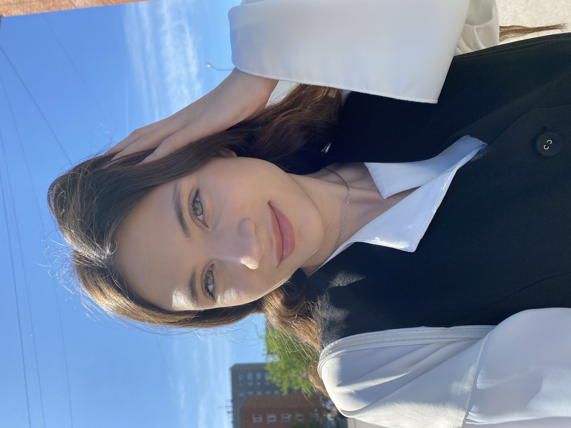A young woman is centered in the frame, capturing a selfie with a slight smile. She is wearing a white shirt with wide sleeves layered under a black vest. A thin silver necklace is visible around her neck. Her brown hair is styled to the side, with one hand gently resting on her head. The background features a clear blue sky and a glimpse of a multi-story brick building with several windows, situated behind some greenery. This photograph was likely taken outside in Votkinsk, Russia.
