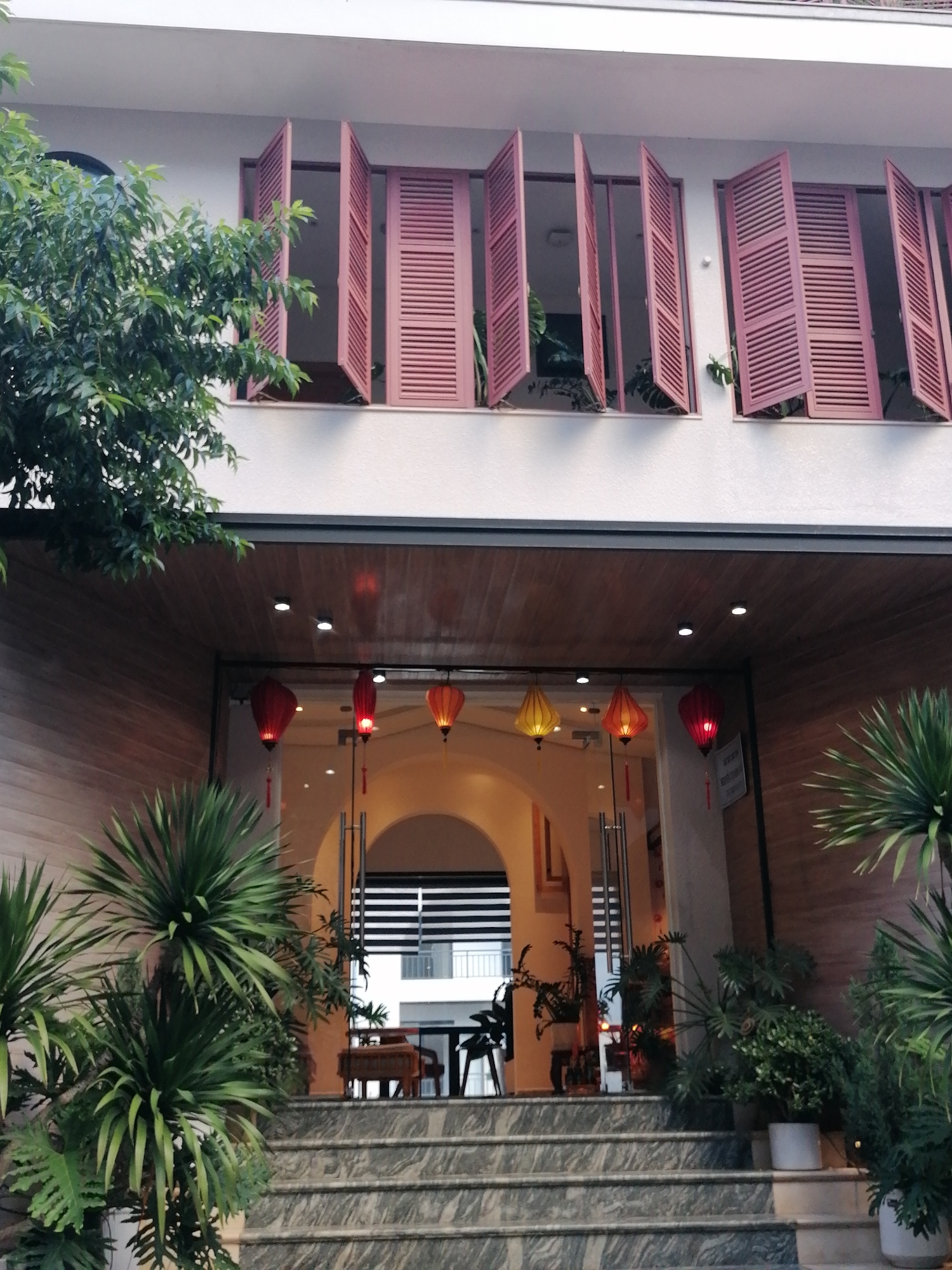The image shows the exterior of a building in Xuân Trường - Đà Lạt, Vietnam. The building has a light-colored facade with several windows on the upper level, each equipped with pink slatted shutters that are partially open. A leafy green tree is visible to the left of the building. The entrance to the building is at ground level, featuring a glass door with a dark frame and a wooden-paneled overhang above. Several traditional Vietnamese lanterns in red, yellow, and orange hang from the overhang. A set of marble-tiled stairs leads up to the entrance, flanked by lush green potted plants. The overall scene suggests a welcoming and aesthetically pleasing environment, likely a hotel or guesthouse.