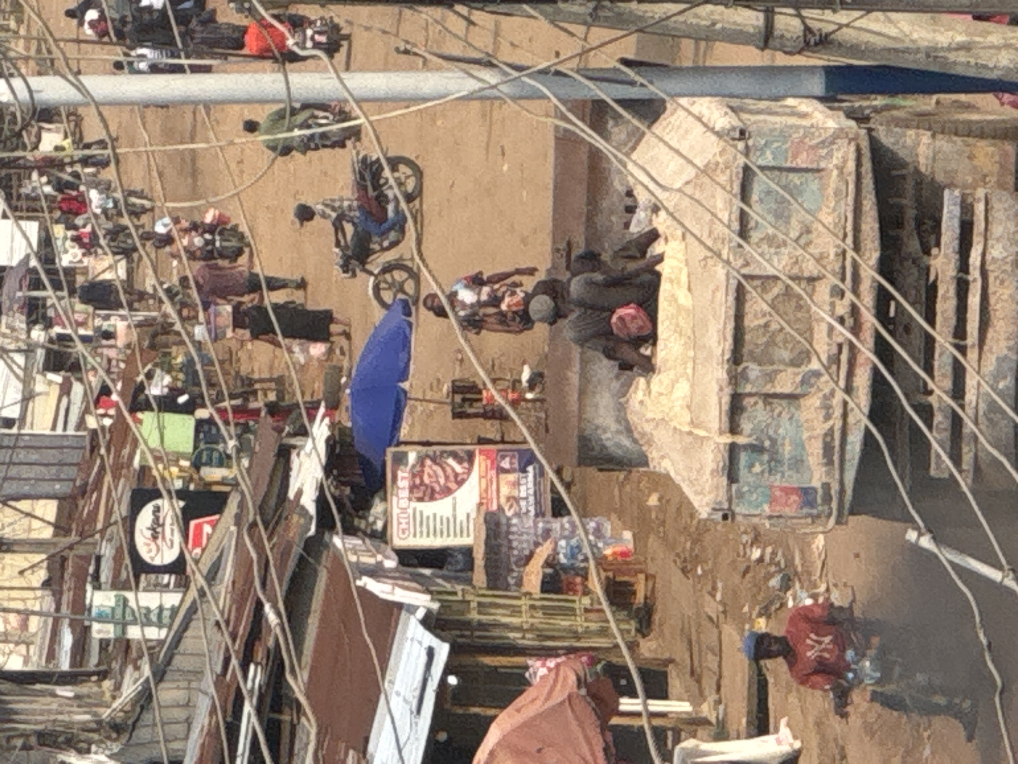 This image captures a bustling outdoor market or street scene in Ihiagwa, Nigeria, characterized by unpaved dirt ground and numerous informal structures that appear to be shops or stalls. The entire frame is crisscrossed by a network of overhead wires, suggesting utility lines.
Many people are present, dressed in casual attire, engaged in various activities. Individuals are seen standing, walking, and gathering in groups. In the mid-left portion, a person is riding a motorcycle, and an open bright blue umbrella rests on the ground. Near this area, market goods are displayed on mats or makeshift tables. A prominent feature on the right side is a large, light-colored dump truck, beside which several men are gathered, some seated. In the bottom-right, a person is visible wearing a red shirt with a white 'X' or cross symbol on the back.
Several signs are visible on the makeshift buildings. One clearly reads "CHIBEST" accompanied by images of food, possibly grilled items, and another adjacent sign has "MWAHH" partly legible. Another sign displays a circular logo, potentially indicating a "BAKERY." The scene is brightly lit, suggesting daytime, and the weather appears clear. The overall impression is one of vibrant daily life and commerce in a community setting.