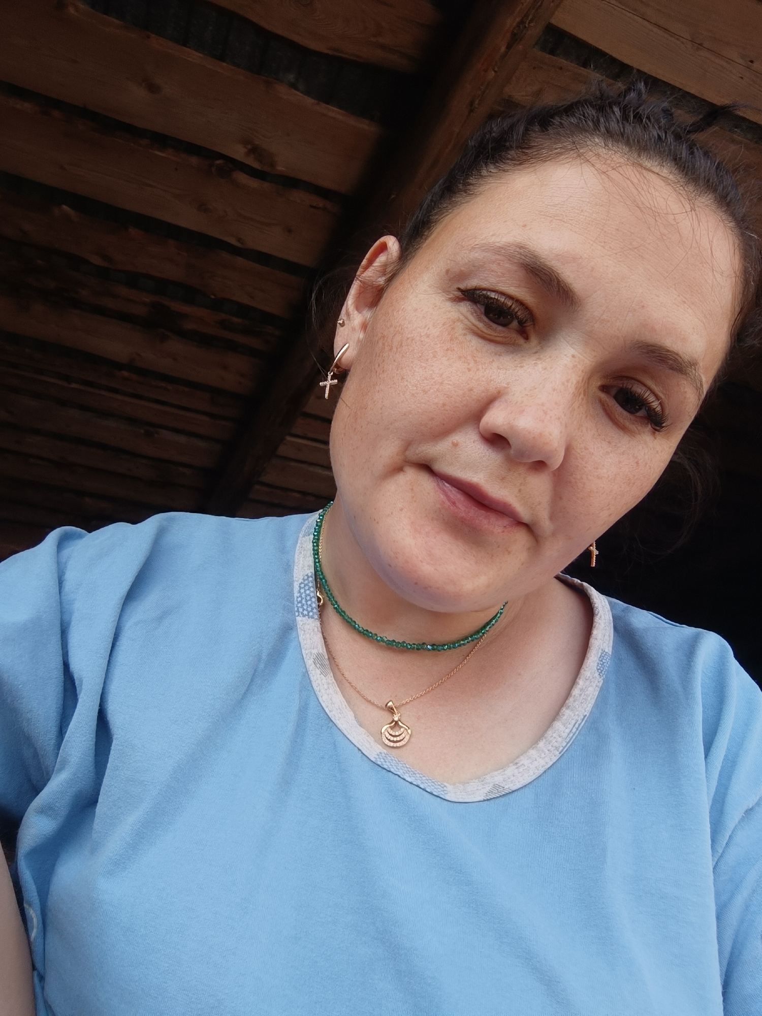 A selfie taken from a low angle of a woman with fair skin and freckles, looking up at the camera. She has her dark hair pulled back and is wearing a light blue t-shirt with a patterned neckline. She is adorned with two necklaces, one a green beaded strand and the other a gold chain with a pendant, and small earrings in her ears. The background consists of weathered wooden planks, suggestive of an outdoor or rustic setting. The image does not contain any information that would allow for a specific location within the city of Karsha, Russia.
