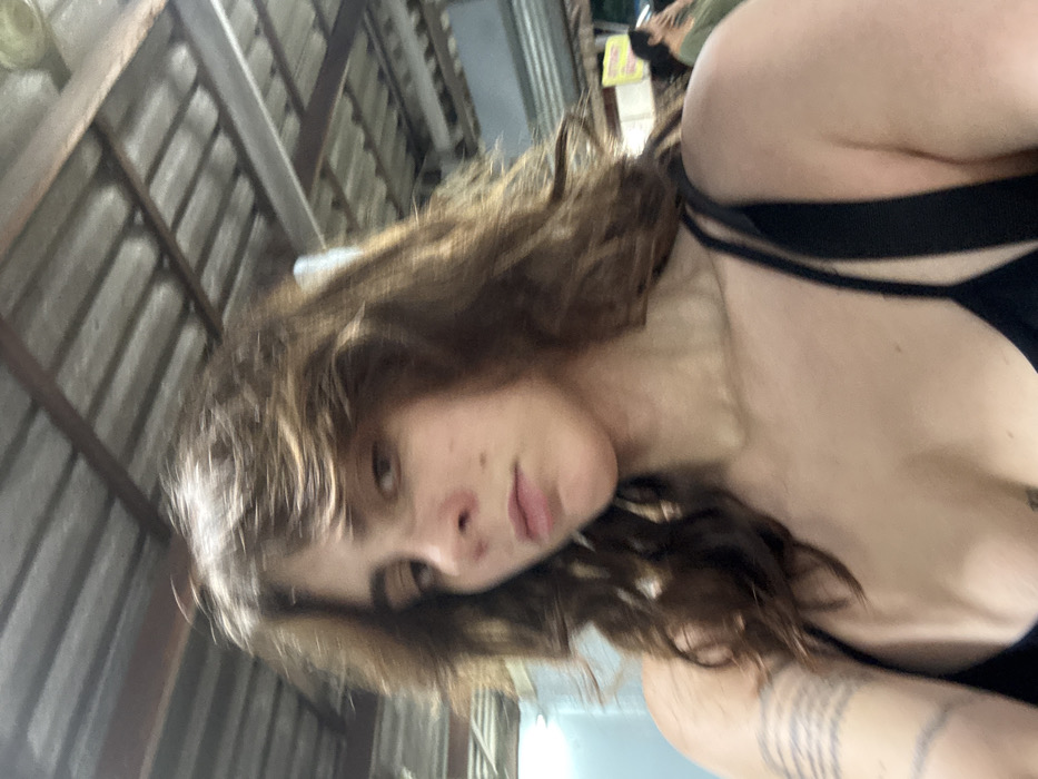 The image presents a close-up, chest-up view of an individual with medium-long wavy brown hair, styled with a middle part, directly facing the camera with a neutral expression. The individual is wearing a black strap top. A tattoo composed of multiple parallel horizontal lines or bands is visible on the left forearm. The background features an overhead structure with a corrugated metal roof supported by exposed beams, suggesting a semi-enclosed outdoor or open-air market-type environment. In the distant right background, a partial view of another person with dark hair and a light-colored top is visible, along with a yellow sign displaying unidentifiable text. The scene is located in the city of Thap Sakae, Thailand.