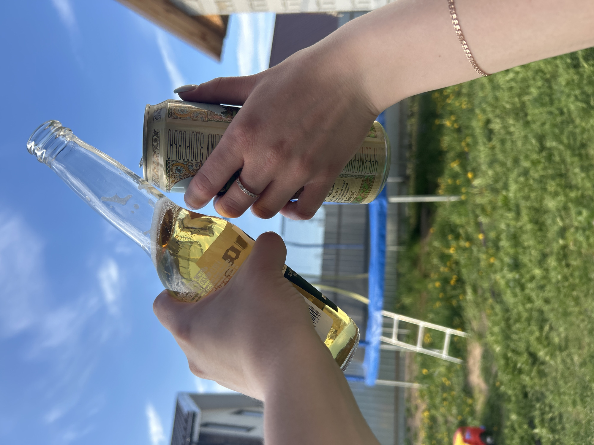A low-angle, outdoor shot shows a person pouring beer from a can into a bottle. A hand with a silver ring is pouring the contents of a green and gold can, which is labelled "ЖИВОЕ ЗДОРОВЬЕ ПРЕМИУМ" in Russian, into a clear glass beer bottle. The label on the bottle is partially obscured. The person pouring wears a gold bracelet. The background includes a trampoline, a ladder, a building, green grass, and a blue sky. The location is Chaikovsky, Russia.