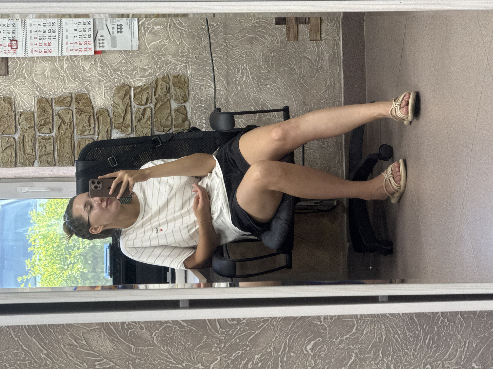 A young woman with her hair in a bun, wearing glasses, a striped t-shirt, and black shorts, sits in a black office chair. She is holding a smartphone in her right hand, taking a selfie in a mirror. Her left hand rests on her lap. She is wearing tan sandals. The background shows a wall with textured wallpaper and a brick pattern, a window with greenery visible outside, and a calendar on the wall. The scene appears to be indoors, possibly in an office or residential space. The floor is tiled. The image is a reflection in a mirror.