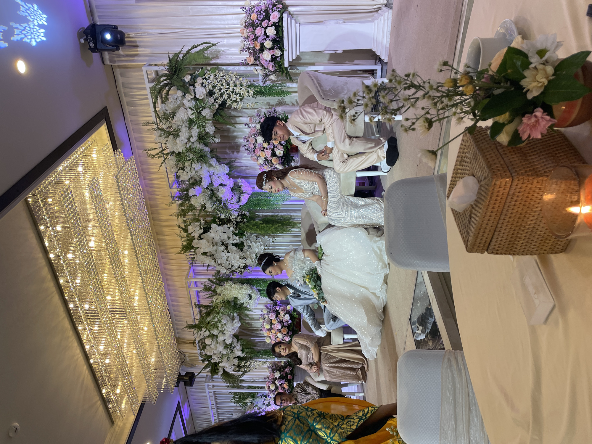 Here is a detailed description of the image:
**Scene:** The image depicts a wedding ceremony and reception.
**Location:** Likely a large event hall or ballroom, presumably in an unknown city, Unknown.
**Environment:** The interior is decorated with a grand chandelier, white drapery, and elaborate floral arrangements.
**People:**
* A bride and groom are the central figures, seated on chairs. They appear to be mid-ceremony or reception.
* Other guests are seated nearby, including women in formal attire.
**Objects:**
* Large crystal chandelier hanging from the ceiling.
* White drapery and floral decorations serve as a backdrop for the ceremony.
* Tables and chairs set for the reception.
* Centerpieces with flowers on the tables.
* A wicker box.
**Actions/Interactions:**
* The bride and groom are the focus of attention, either during the ceremony or receiving congratulations from guests.
* Guests are seated, presumably watching the ceremony or waiting for the reception to proceed.
**Details:**
* The bride is wearing a white wedding gown.
* The groom is wearing a suit.
* Floral arrangements are in white and pastel shades.
* The lighting is soft and romantic.