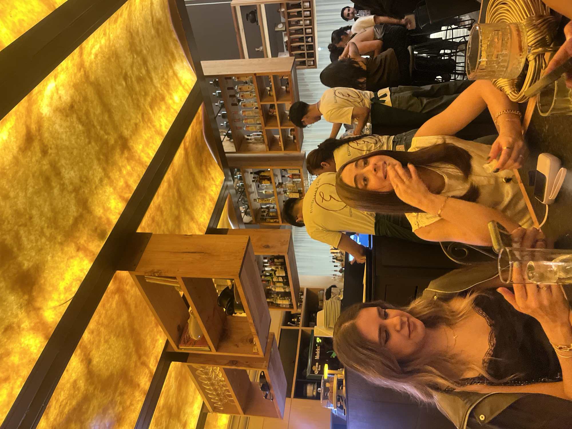 The image depicts the interior of a bar in San Andrés Cholula, Mexico. The scene is populated with several individuals. Two women are prominently positioned in the foreground. One woman is wearing a black top with lace detailing and a green jacket. The other woman wears a white top. They appear to be seated at a counter. In the background, other people are visible, including staff wearing shirts with the logo of "ELUVIER'S BISTRO & CAFÉ". The staff are tending to the bar, which is lined with shelves stocked with bottles of liquor and glassware. Beer taps with Corona and Modelo logos are visible. The bar has a distinctive design with a patterned yellow ceiling and wooden shelving units.