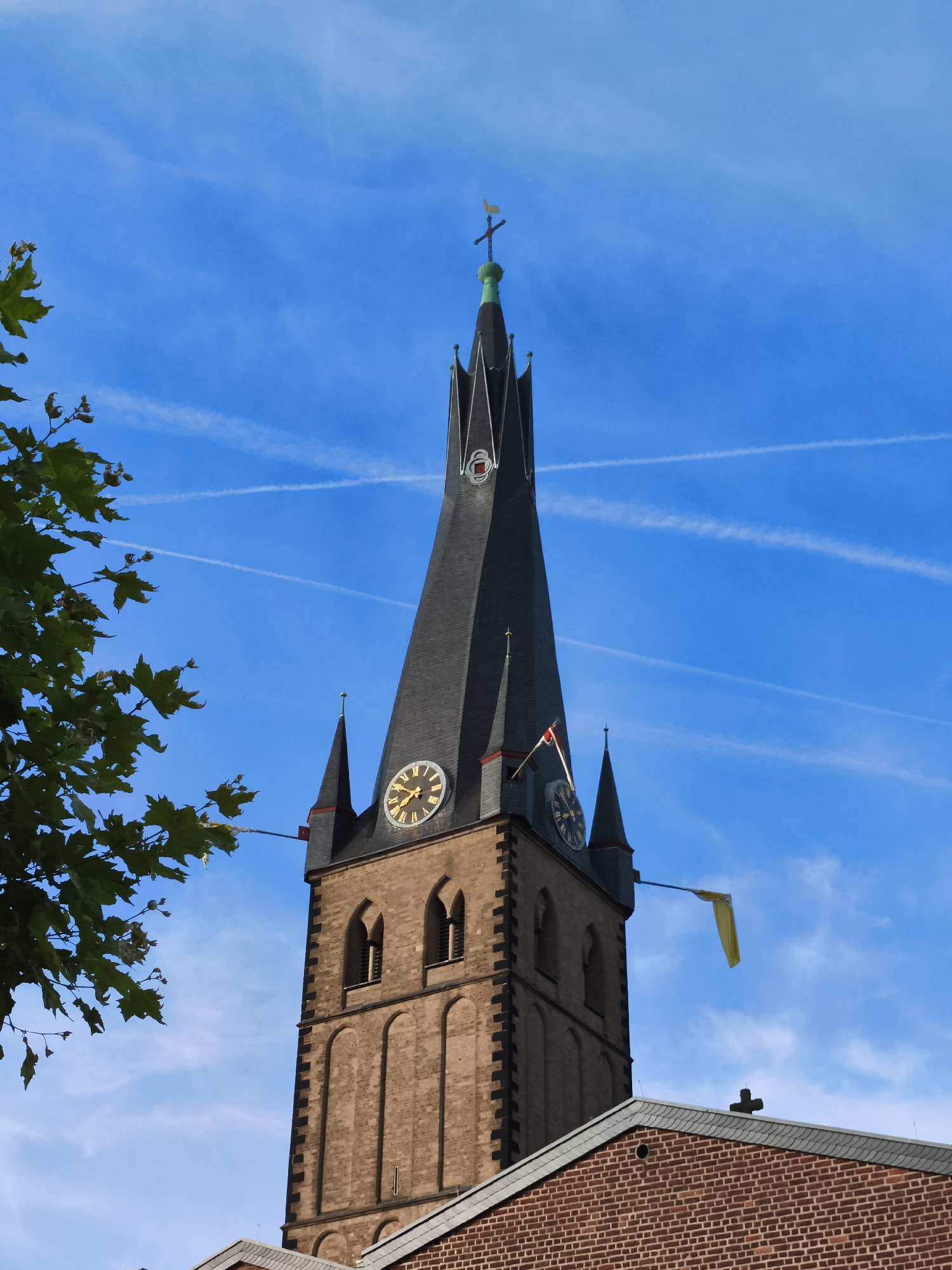 This image depicts the spire of a church against a bright blue sky with wispy clouds and airplane contrails. The spire is made of dark grey material and tapers to a point, topped with a cross and a weather vane. Two clock faces are visible on the tower, one at approximately 10:10 and another at approximately 2:40. A small yellow flag flies from a flagpole attached to the tower. To the left of the spire, a portion of a leafy tree is visible. The lower part of the image shows a section of a brick building with a slate roof, also topped with a cross. The architecture suggests a European setting, and the context provided identifies the location as Dusseldorf, Germany. The clear sky and bright sunlight indicate it is daytime. No people are visible in the image.