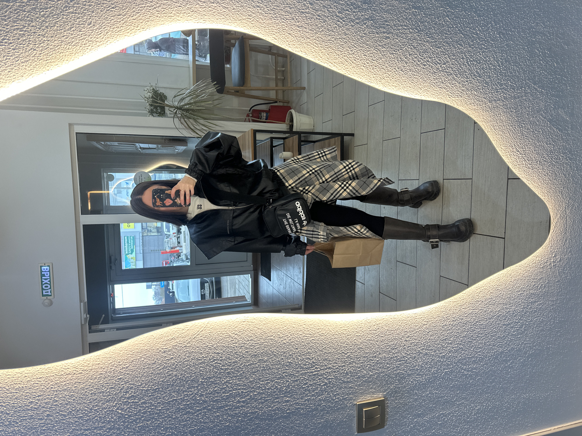 A person is reflected in an illuminated mirror. The individual is wearing a black jacket, a plaid skirt, leggings, and knee-high boots. They are holding a brown paper bag. The location appears to be inside a building, possibly a shop or restaurant, as indicated by the interior decor and the presence of an exit sign in Russian ("ВРІХОП"). Visible through the glass door are cars and buildings. The reflection seems to show a street scene, indicating a possible location in the city of Votkinsk, Russia, based on the Russian text on the exit sign. The individual is holding a smartphone up in front of their face, likely taking the picture.