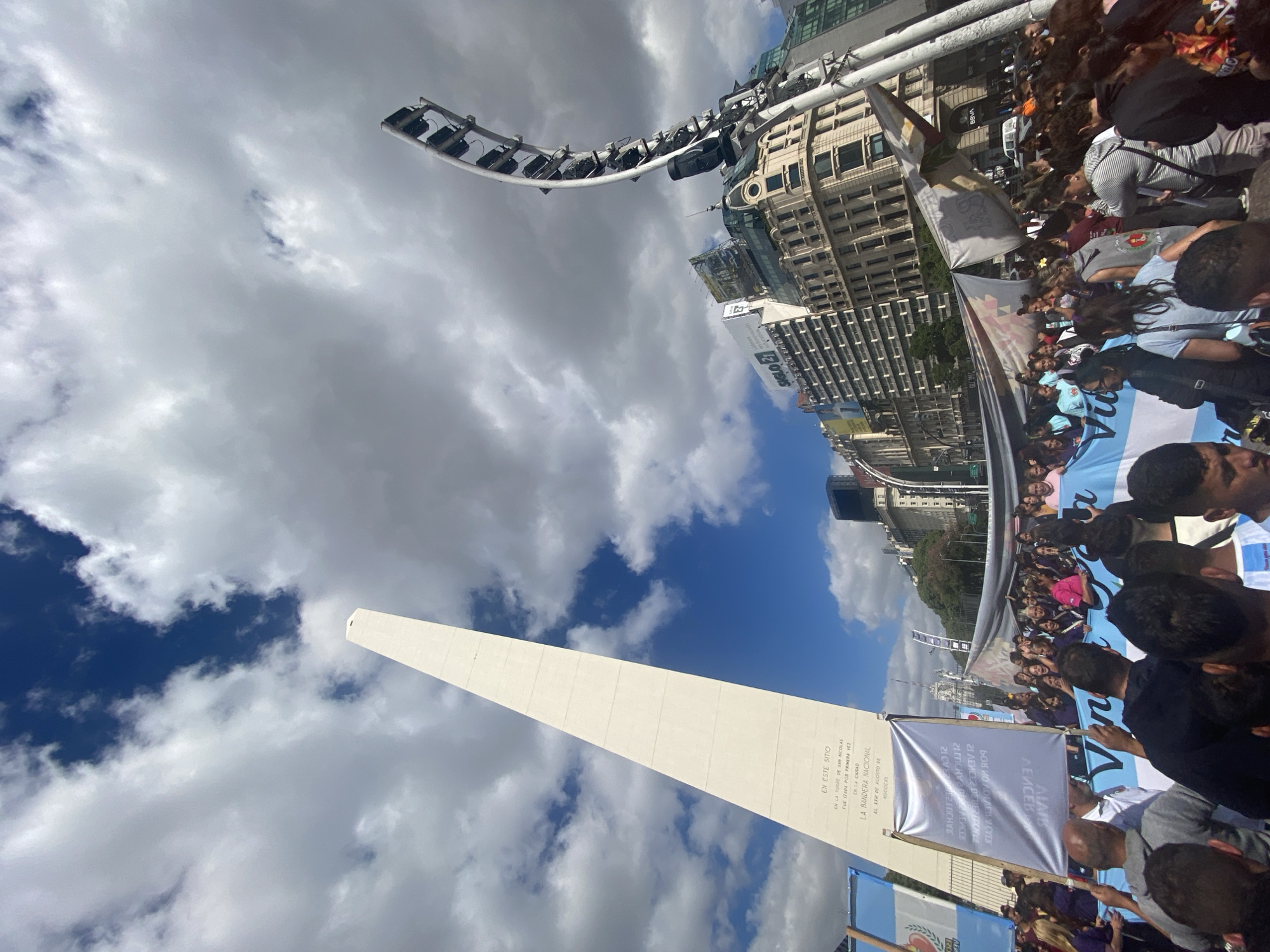 The image shows a street-level view of the Obelisco, a historical monument in the city of Buenos Aires, Argentina. It is a white obelisk under a sky partially covered with white, puffy clouds. A crowd of people is gathered around the base of the obelisk. Many are holding banners and Argentinian flags. There are buildings visible in the background.