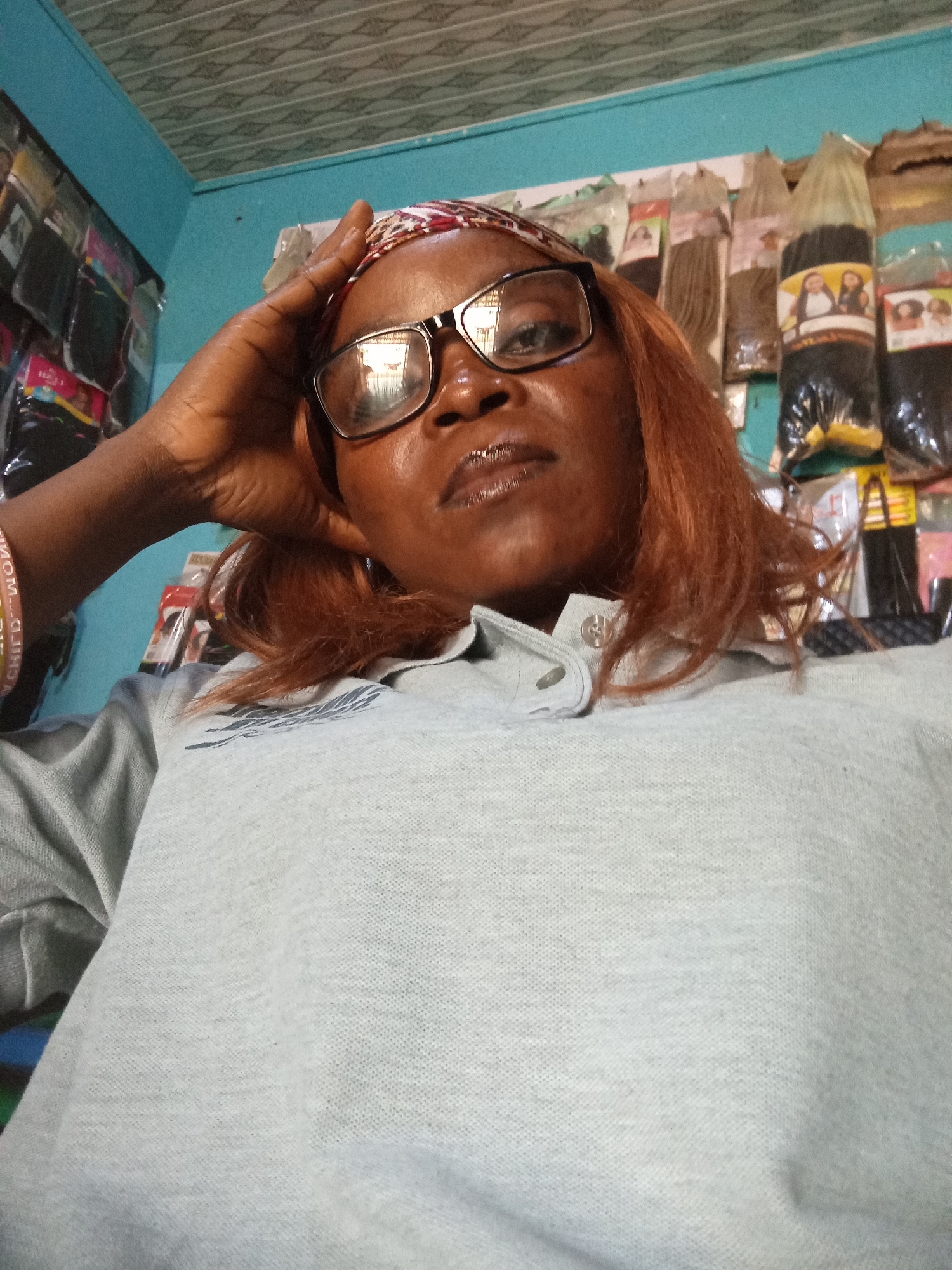 A middle-aged Black woman with reddish-brown hair, wearing glasses and a light blue polo shirt, is looking directly at the camera. Her right hand is on her forehead, and she is wearing a patterned headband. Behind her, hair extensions are displayed on racks in what appears to be a salon or shop. The walls are painted light blue, and the ceiling is paneled. The image was taken in Jalingo, Nigeria.