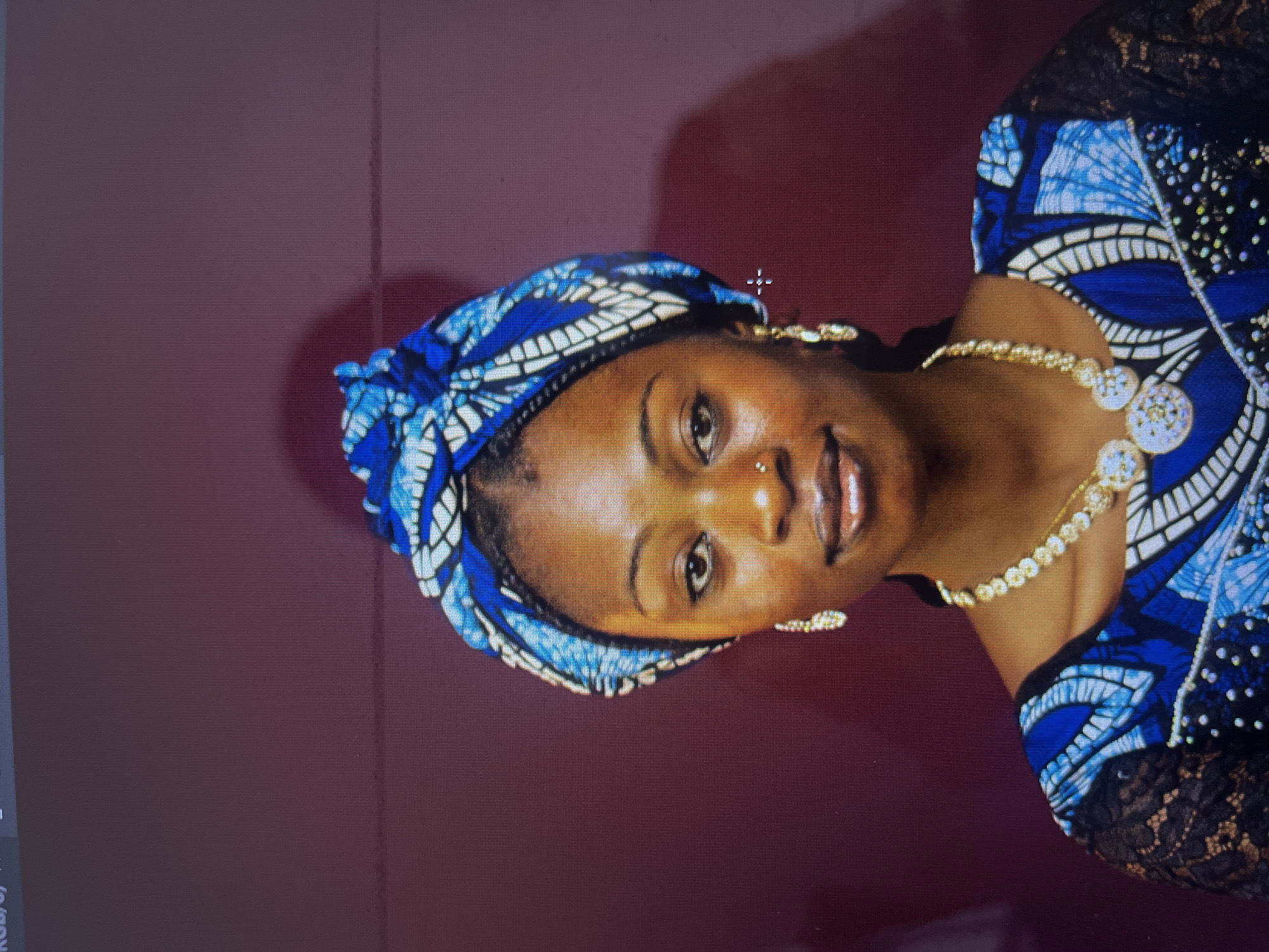 The image is a headshot of a young Black woman wearing a blue and white patterned headwrap and a matching blue dress with black lace sleeves. She is adorned with a gold necklace with large circular pendants and dangling earrings. A small nose stud is also visible. She has dark eyes, full lips, and her skin appears smooth. The background is a dark, reddish-purple wall with a faint shadow of a person on the right side. The image is likely a studio portrait, taken indoors. There is no discernible information within the image that would indicate its specific location in Gura Top, Nigeria.