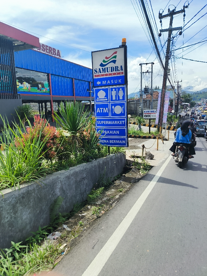 A blue sign with white lettering advertises the Samudra Serba shopping center at a street intersection in Ciwidey, Indonesia. The sign is mounted on a metal pole and is surrounded by a concrete planter with green grass and plants. The sign's lettering states "Masuk," "ATM," "Supermarket," "Pakaian," and "Arena Bermain." There is a white painted line separating the road from a sidewalk where a man wearing a blue jacket and a black helmet is driving a motorcycle. In the distance, there is a two-lane paved road with several vehicles traveling on it. The road is lined with trees on the opposite side. The sky is blue with white clouds and the weather appears sunny. The setting is likely an urban or suburban area based on the presence of the shopping center, road, and surrounding buildings. The scene is a typical Indonesian street environment, with traffic, pedestrians, and businesses present. The image's subject is the Samudra Serba shopping center sign, providing information about the amenities available at the location.