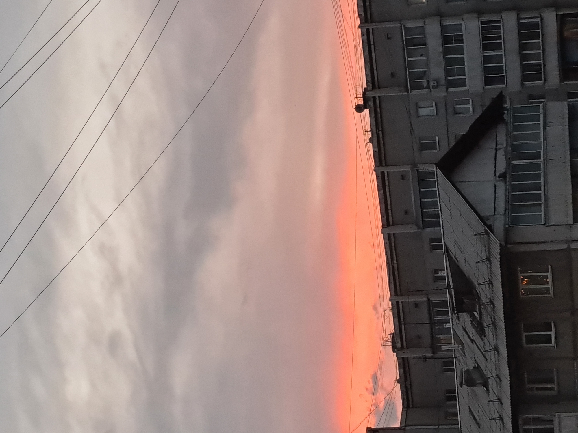 The image shows a view of apartment buildings against a twilight sky in Irkutsk, Russia. The sky is colored with hues of orange and pink, indicating a sunset. Power lines are visible against the sky. The buildings are constructed from concrete and have multiple stories with many windows. One building in the foreground has a sloped, corrugated metal roof with chimneys. The overall impression is of a typical urban residential area at dusk.