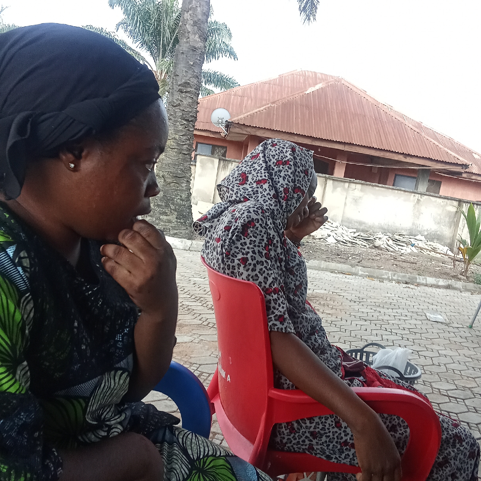 Two women sit outdoors on plastic chairs. One woman wears a black head covering and a green and black patterned dress, her hand is raised to her chin. The other woman wears a grey patterned head covering and dress, with red accents. They are sitting on a paved surface near a building with a brown corrugated metal roof. A palm tree is visible in the background. In Akwanga, Nigeria.