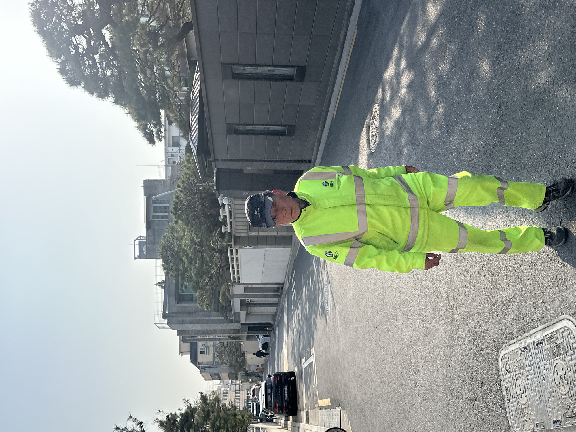 An adult male, appearing middle-aged or older, walks directly towards the viewer on a paved asphalt street in Seoul, South Korea, specifically within the Seodaemun-gu district. He occupies the foreground, slightly right of center. He is dressed in a neon yellow two-piece uniform featuring horizontal grey reflective stripes on the chest, arms, and legs. On his left chest, a logo is visible with the Korean text "서대문구" (Seodaemun-gu). He wears a dark grey baseball cap with a white "eider" logo on the front, and dark athletic shoes.
The street is lined on both sides by contemporary multi-story buildings, some with concrete facades and flat roofs, and others with dark grey perimeter walls. Large, manicured pine trees are interspersed among the structures. On the left side of the street, multiple vehicles are parked, including at least two black crossovers; one is a black Mini Cooper Countryman displaying the Korean license plate "157누 9044". A light grey, rectangular manhole cover with a geometric pattern is present on the asphalt in the left foreground, and a circular manhole cover is visible further down the road. Another silhouetted figure walks in the distance on the left side of the street. The scene is illuminated by bright sunlight under a clear, light blue sky, casting distinct shadows from trees and buildings across the roadway.