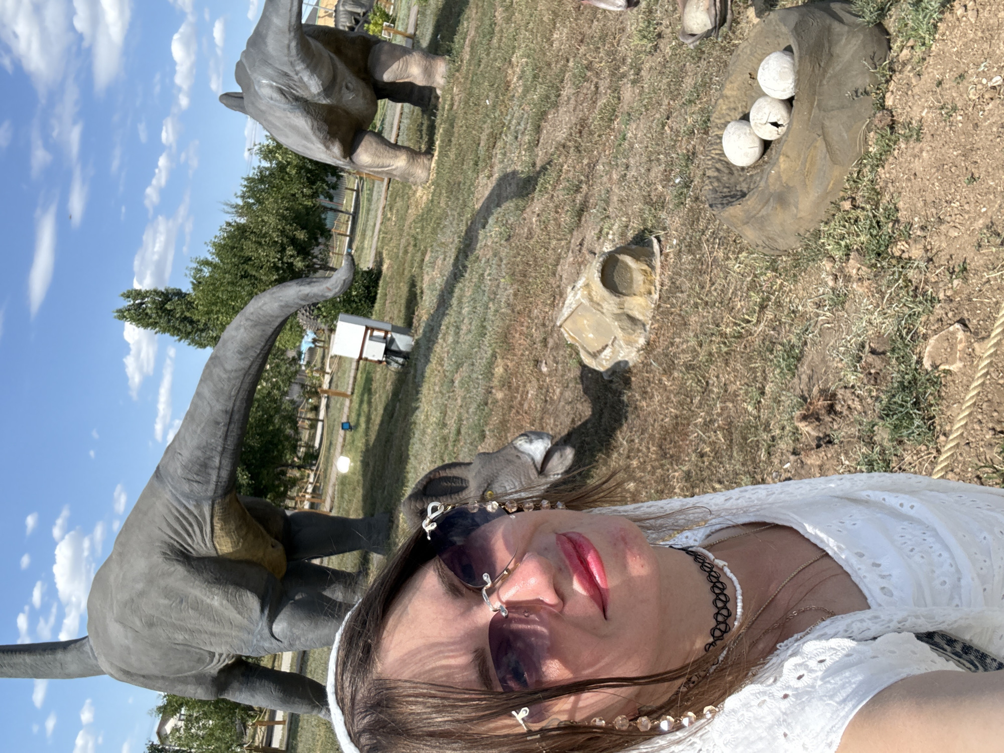 A woman is taking a selfie with a dinosaur display in the background. The woman is wearing a white dress, sunglasses, and jewelry. She is smiling at the camera. In the background, there are several large dinosaur statues, including one that appears to be a brontosaurus. There are also some dinosaur eggs in a nest on the ground. The scene is set in a park-like environment with grass and trees. The sky is blue with some clouds. This appears to be a tourist attraction in the city of Sol-Iletsk, Russia.
