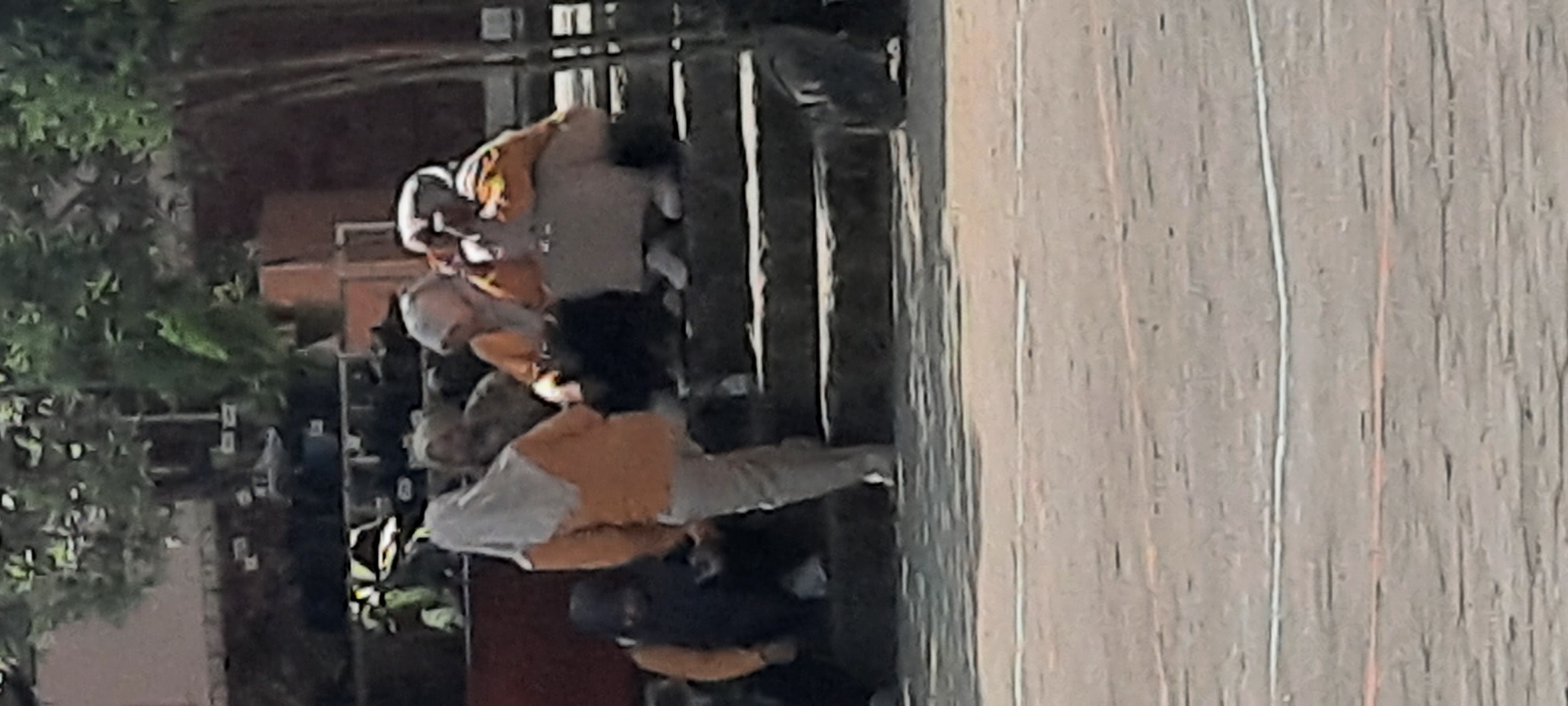 In Tangkil, Indonesia, a group of individuals are gathered on a set of outdoor stairs. The individuals, likely students given their attire, are wearing headscarves and loose-fitting clothing. One person in the foreground, on the left, is wearing a dark headscarf and an orange top. To their right, another person wears a light grey headscarf and an orange top, with grey pants. Further to the right, two people sit on the stairs; one in an orange top with a light-colored headscarf, and another in an orange top and a light-colored headscarf. A dark object, possibly a backpack, is situated on the stairs to the far right. The setting appears to be a school or a similar institution, with trees and greenery visible in the background, and a paved area in the foreground. The stairs themselves are made of dark stone or concrete.