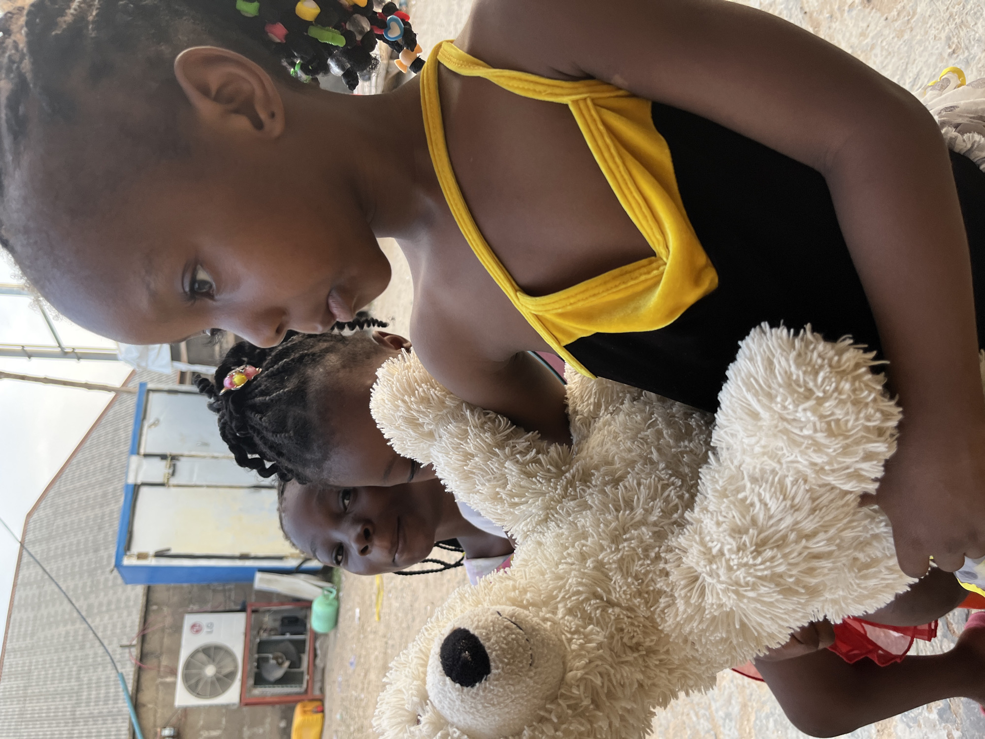 Three young girls are in close proximity, with one holding a large, cream-colored teddy bear. The foremost girl wears a black top with yellow straps and has braided hair adorned with colorful beads. The second girl behind her has braided hair with a single flower accessory. The third girl is partially visible, her expression visible. The background includes a building with an air conditioning unit and a blue and white structure above. The location appears to be Iddah, Nigeria.