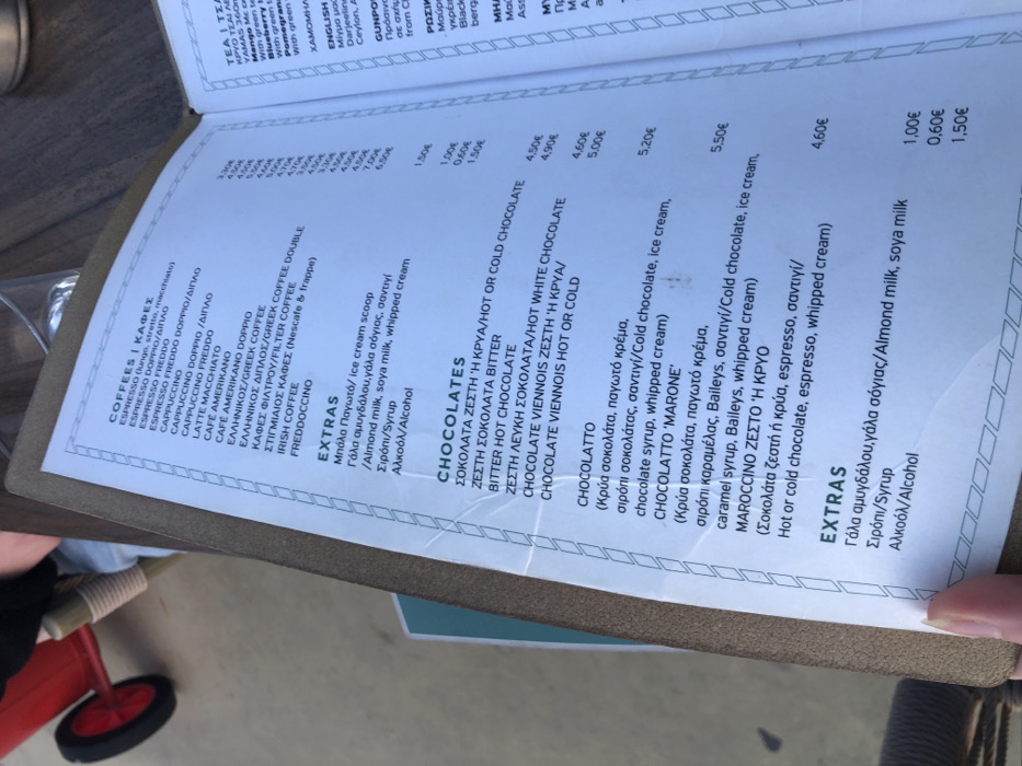 A paper menu on a brown wooden table, listing the prices for coffee and chocolate drinks in a cafe located at 37.929, 23.685 in Palaio Faliro, Greece. The menu is in Greek. The paper is folded in the upper left corner, revealing a red toy wagon partially visible underneath. The camera is pointing directly at the menu. The table is outdoors. The menu lists the cost of various types of coffee, including espresso, cappuccino, latte, americano, and freddo cappuccino. It also lists the cost of different types of chocolate drinks, including hot chocolate, white chocolate, bitter chocolate, and "Marone." The drinks are available hot or cold. There is also a list of extra ingredients that can be added to the drinks, including milk, cream, ice cream, and whipped cream. The prices range from 1.00€ to 5.50€.