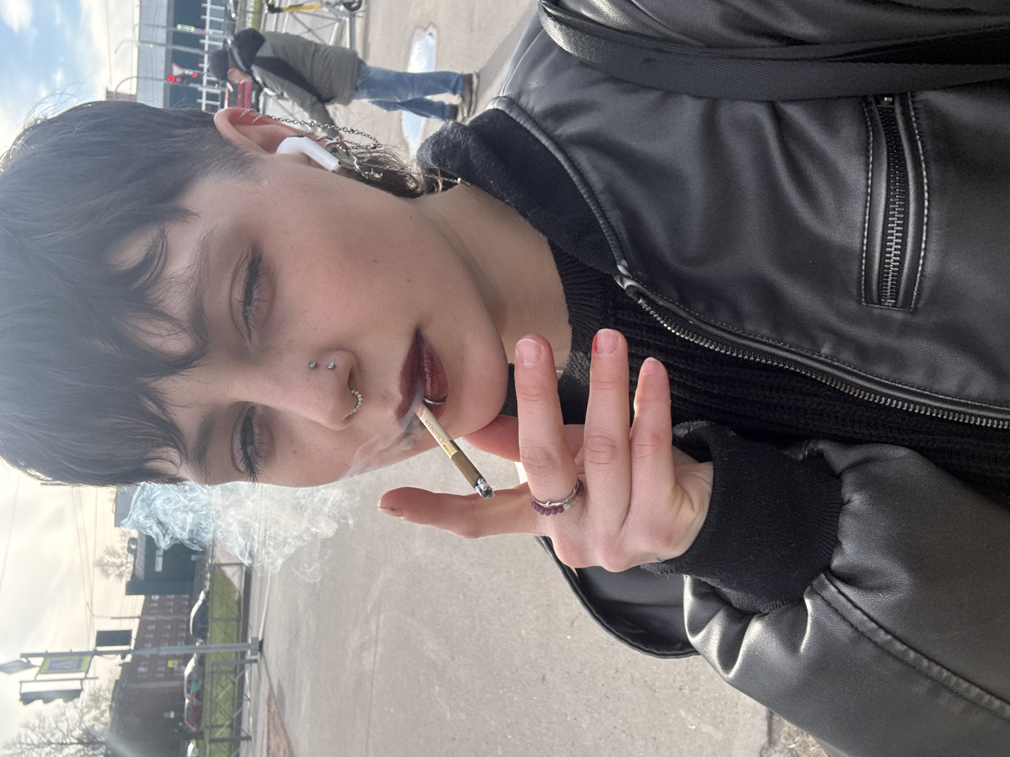 A person with short dark hair, multiple piercings in their left ear and nose, and wearing a black leather jacket and a black sweater, is smoking a cigarette outdoors. They are holding the cigarette between their fingers, and smoke is visibly rising from the lit end. The person is wearing a ring on one of their fingers. In the background, there is a street scene with traffic lights, buildings, and other people. The location appears to be Saint Petersburg, Russia, based on the architectural style of the buildings and the traffic signals.